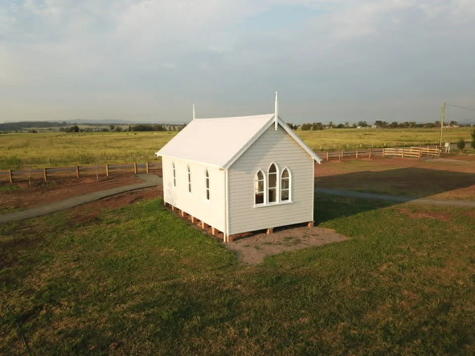Small white church with stained glass windows, set on wooden stilts, in a rural landscape with green grass and open fields.