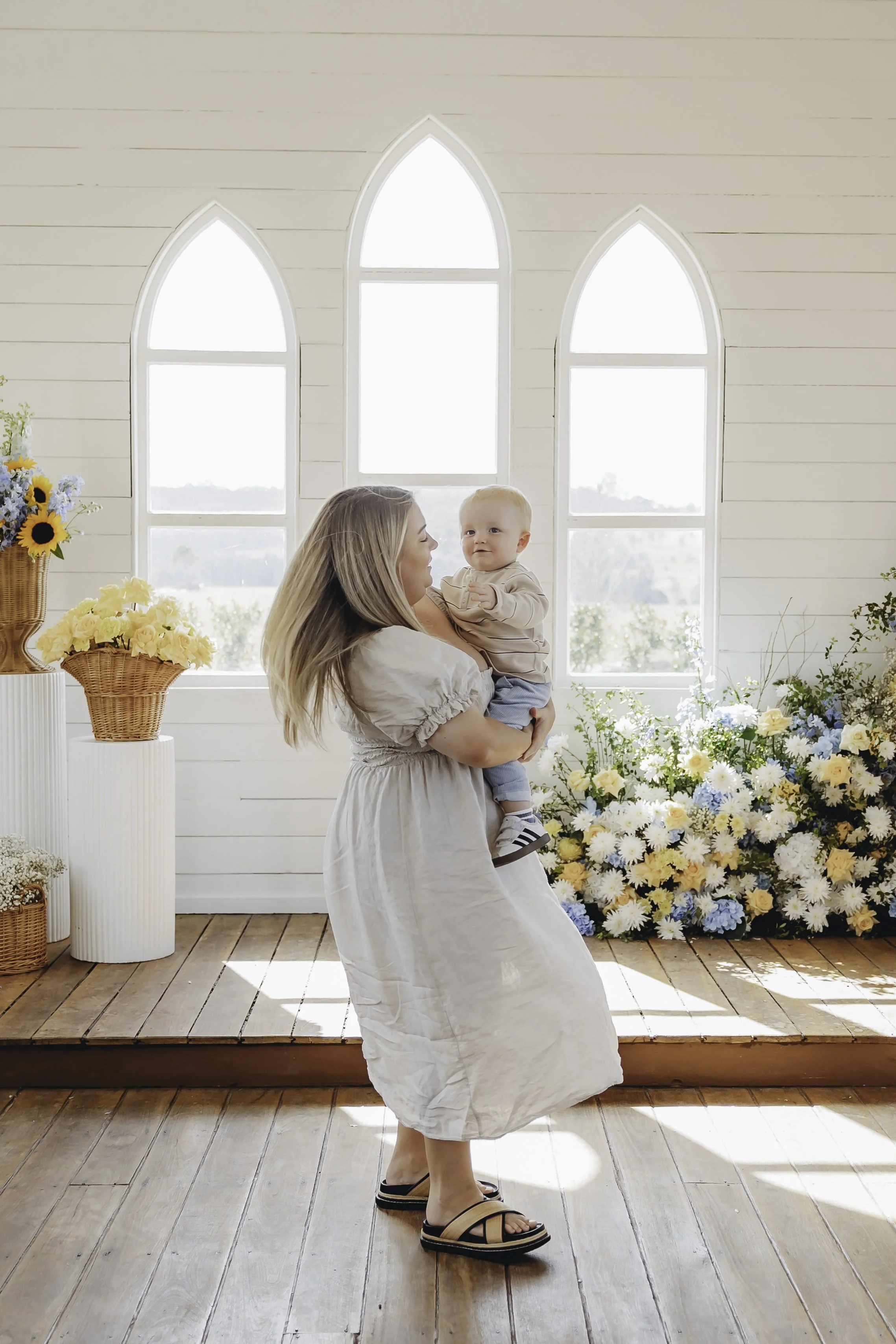 A woman holding a baby inside a bright room with high arched windows, surrounded by flower arrangements and baskets of flowers on pedestals.