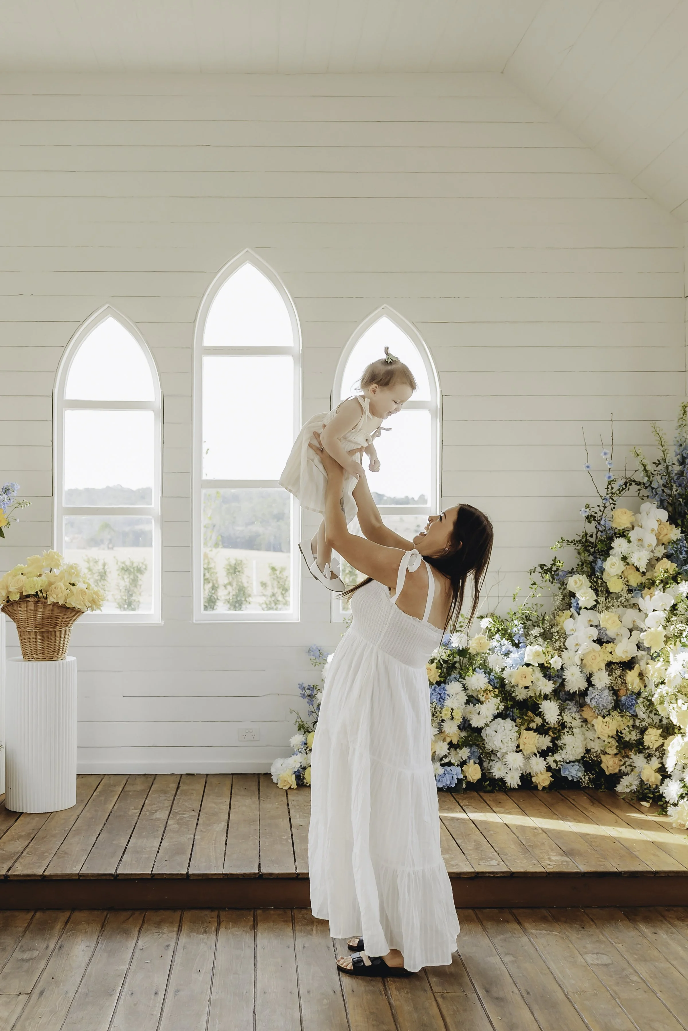 A woman in a white dress lifting a young girl in a cream-colored dress in a bright room with large arched windows and floral arrangements.