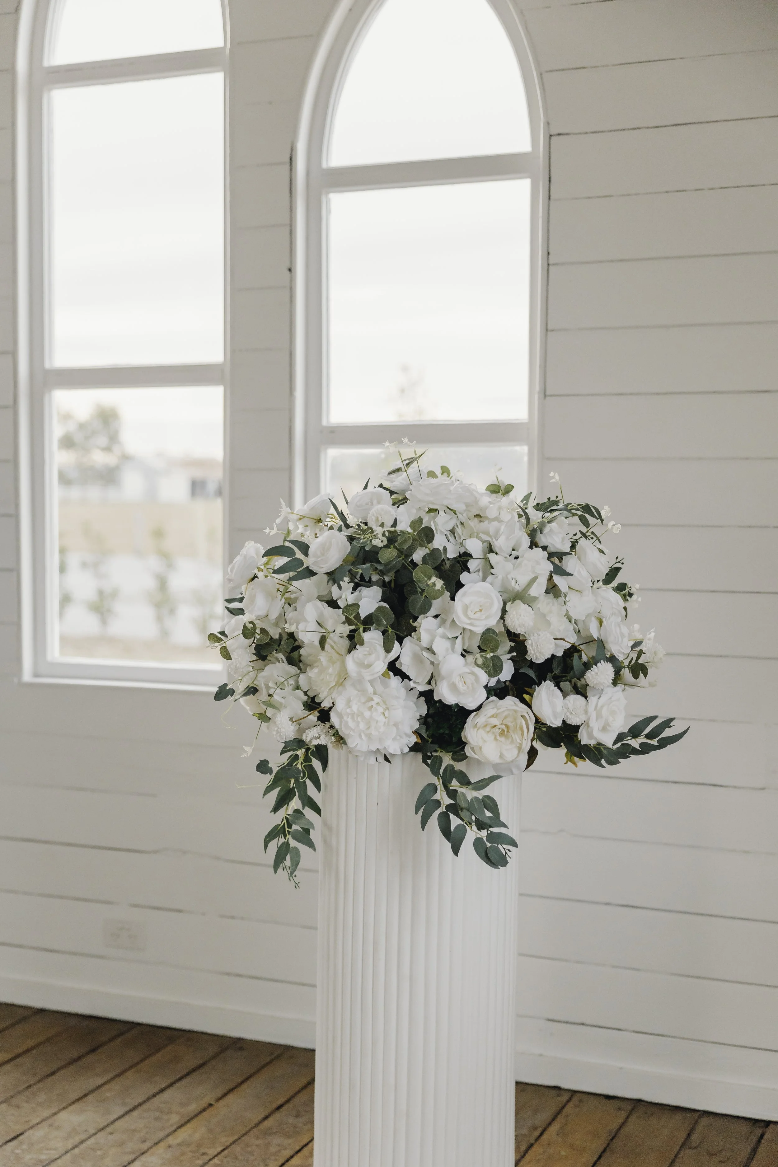 A bouquet of white flowers with greenery in a white vase, placed on a white fluted pedestal in front of white shiplap walls and windows with curved tops.