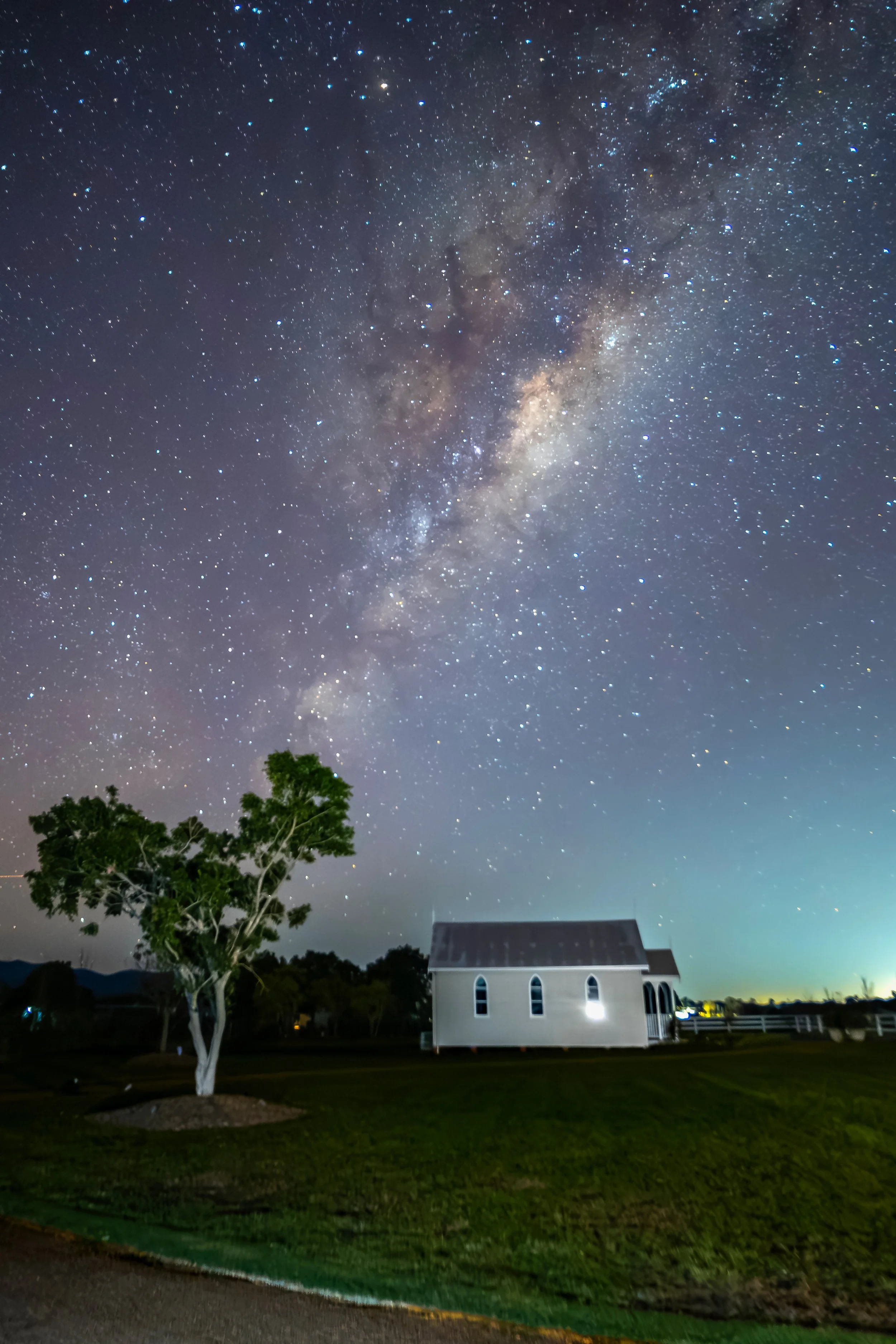 Night sky filled with countless stars and the bright band of the Milky Way galaxy. A white church with arched windows and a gabled roof is in the foreground, with a tree nearby and a dark landscape beyond.