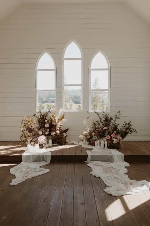 Interior of a white chapel-style room with three arched windows, decorated with large floral arrangements on white cloth-covered tables on the wooden floor.