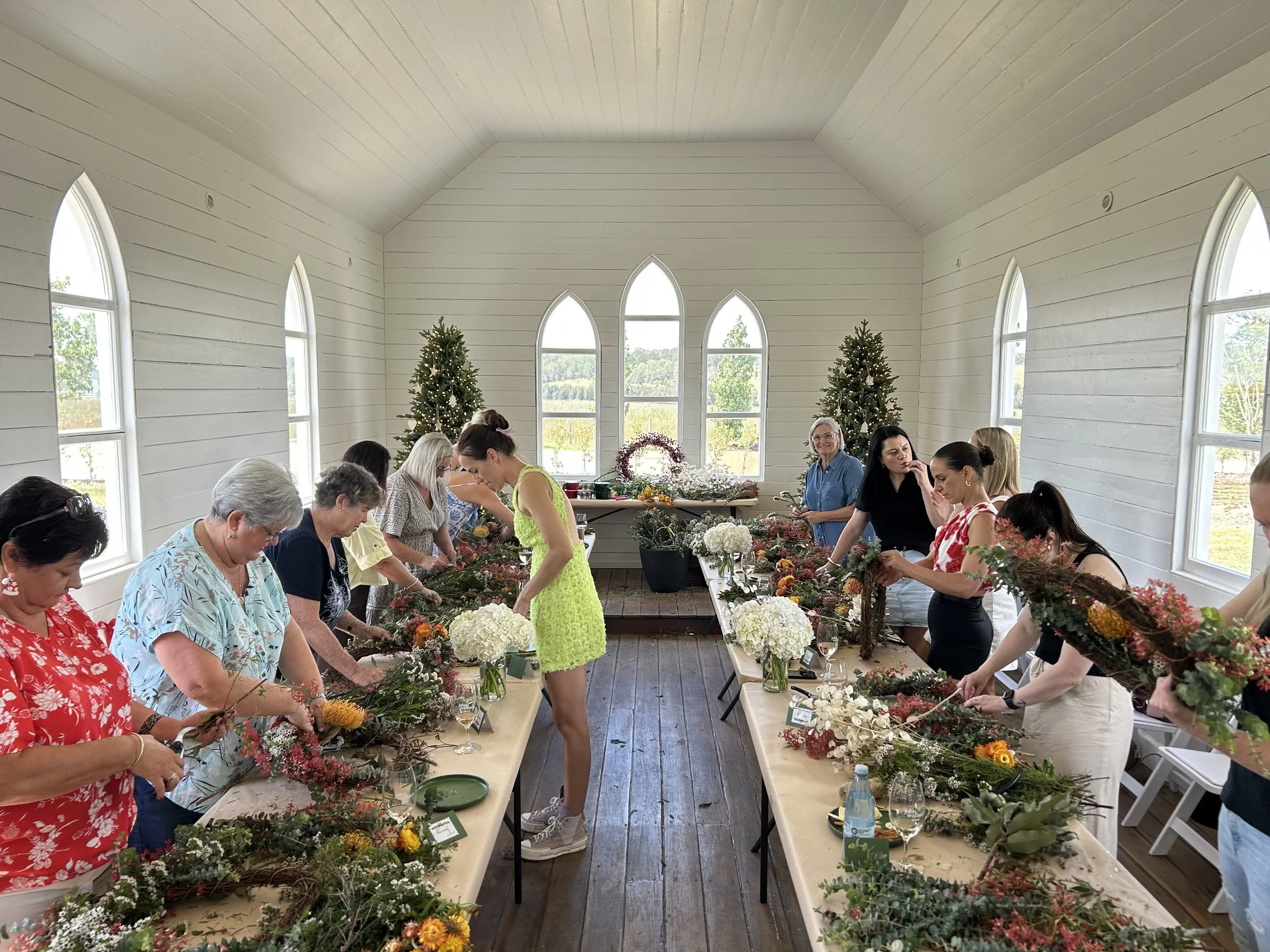 Women participating in a floral arrangement workshop in a bright room with white wooden walls and arched windows, decorated with Christmas trees.
