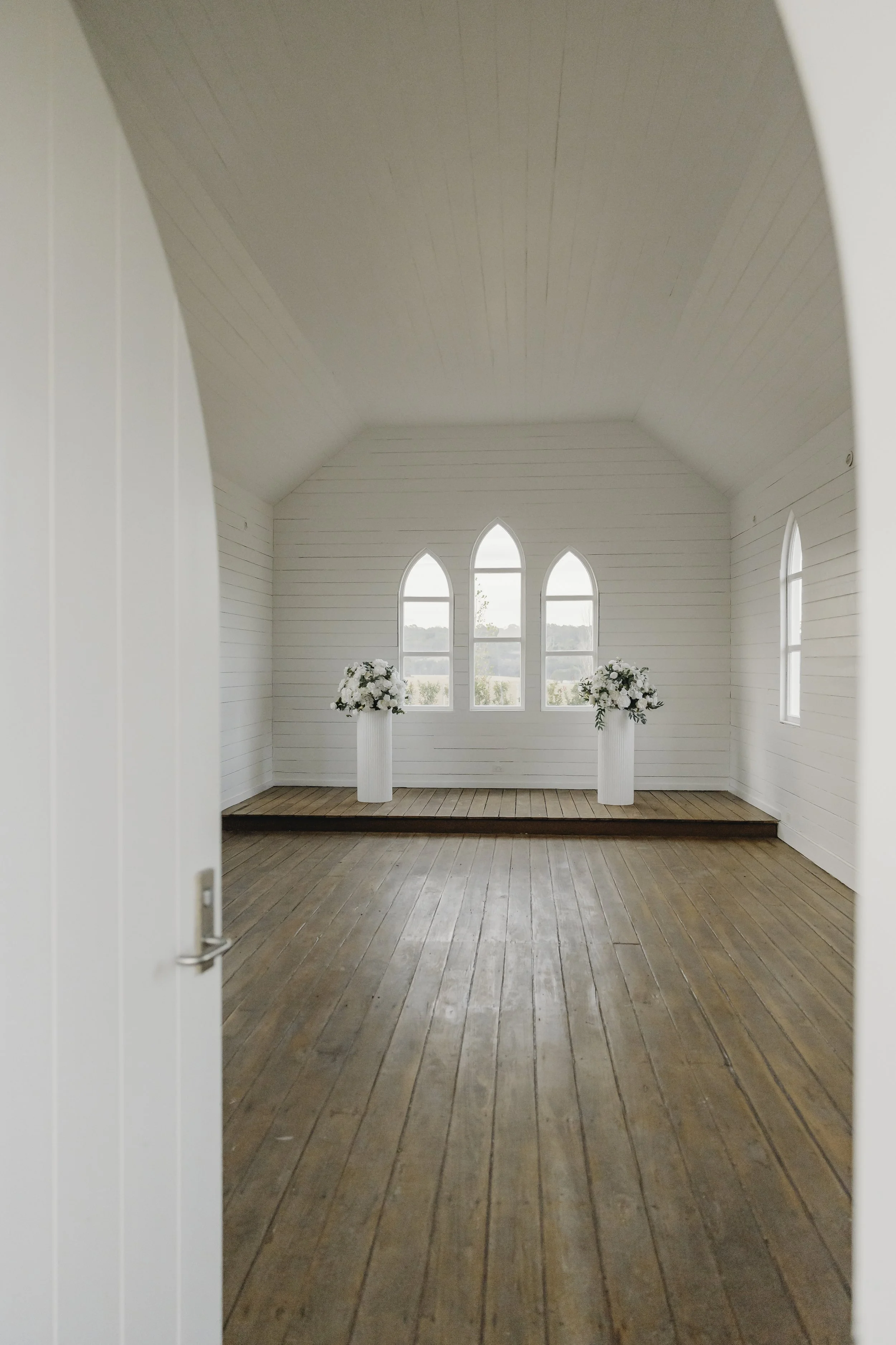 View of the front of a white wedding chapel with three arched windows and two white columns with floral arrangements.