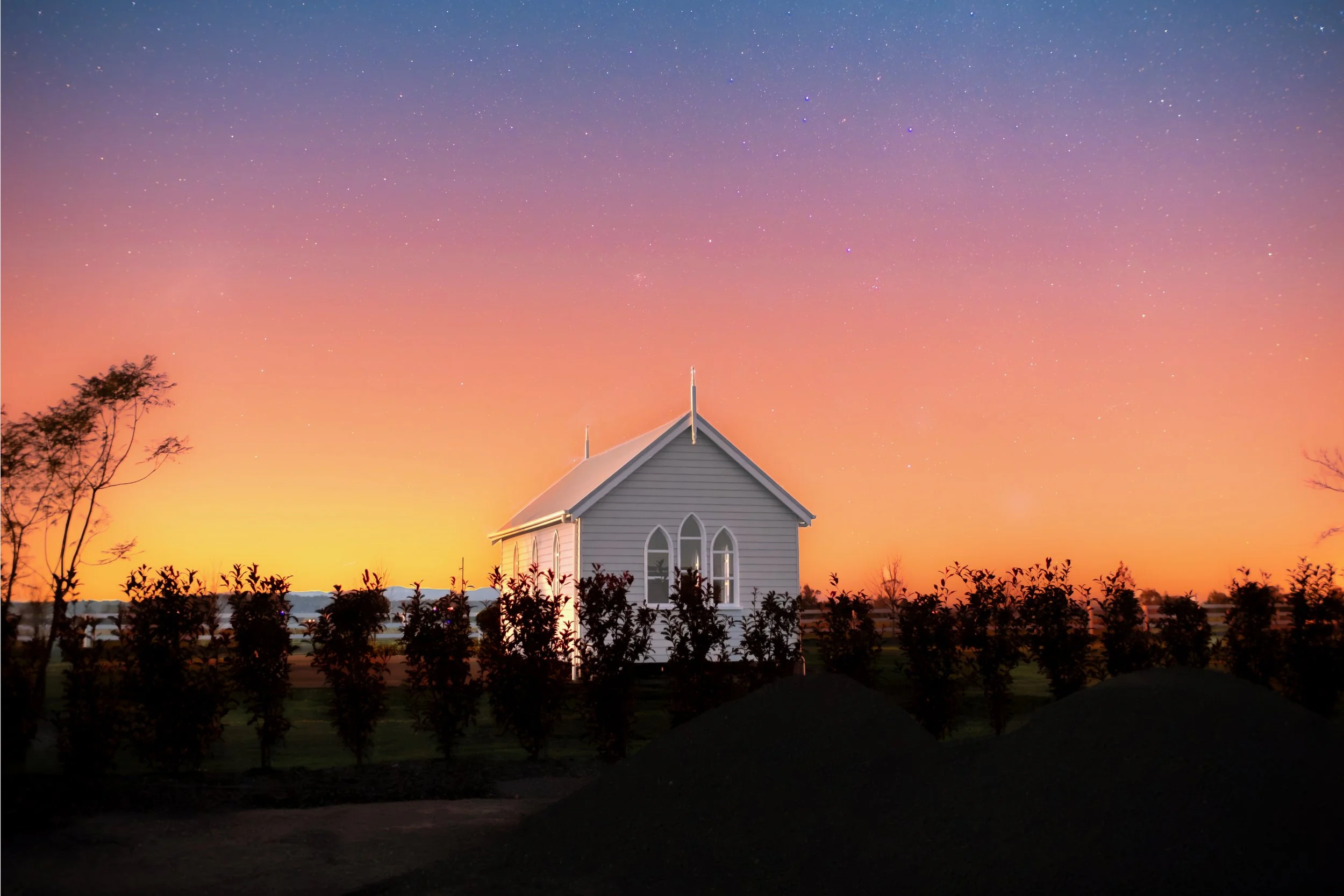 A small white church with pointed windows beneath a colorful sunset sky filled with countless stars.