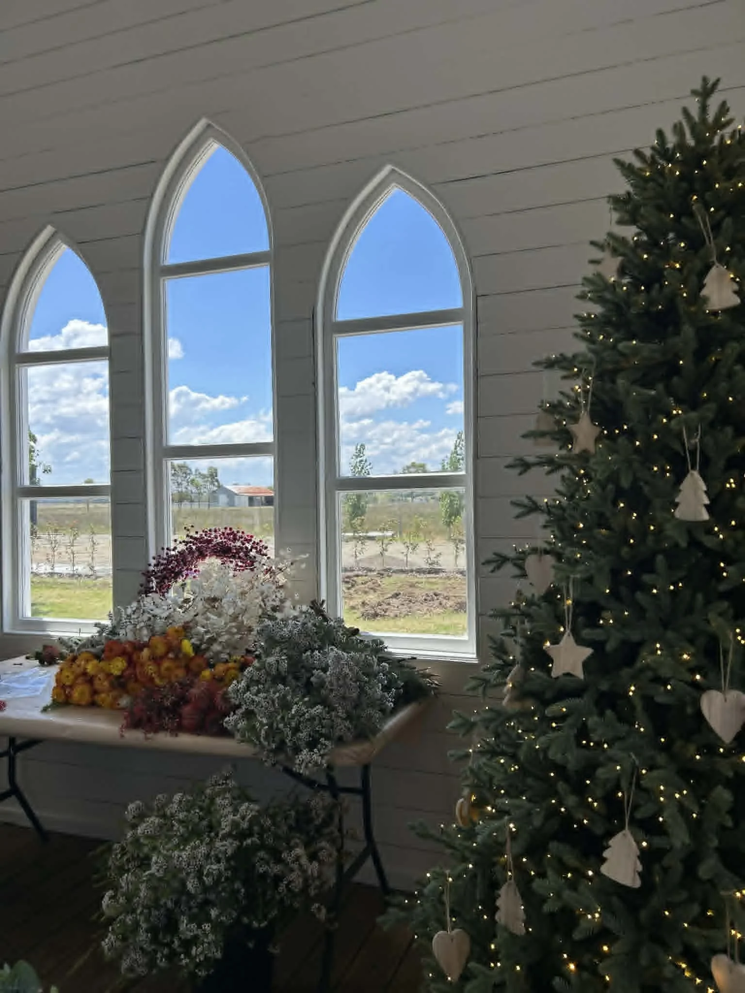 Interior of a room with three large, pointed arch windows showing blue sky and white clouds outside. Inside, a decorated Christmas tree with lights and star ornaments stands next to a table with a flower arrangement and other decorative items.