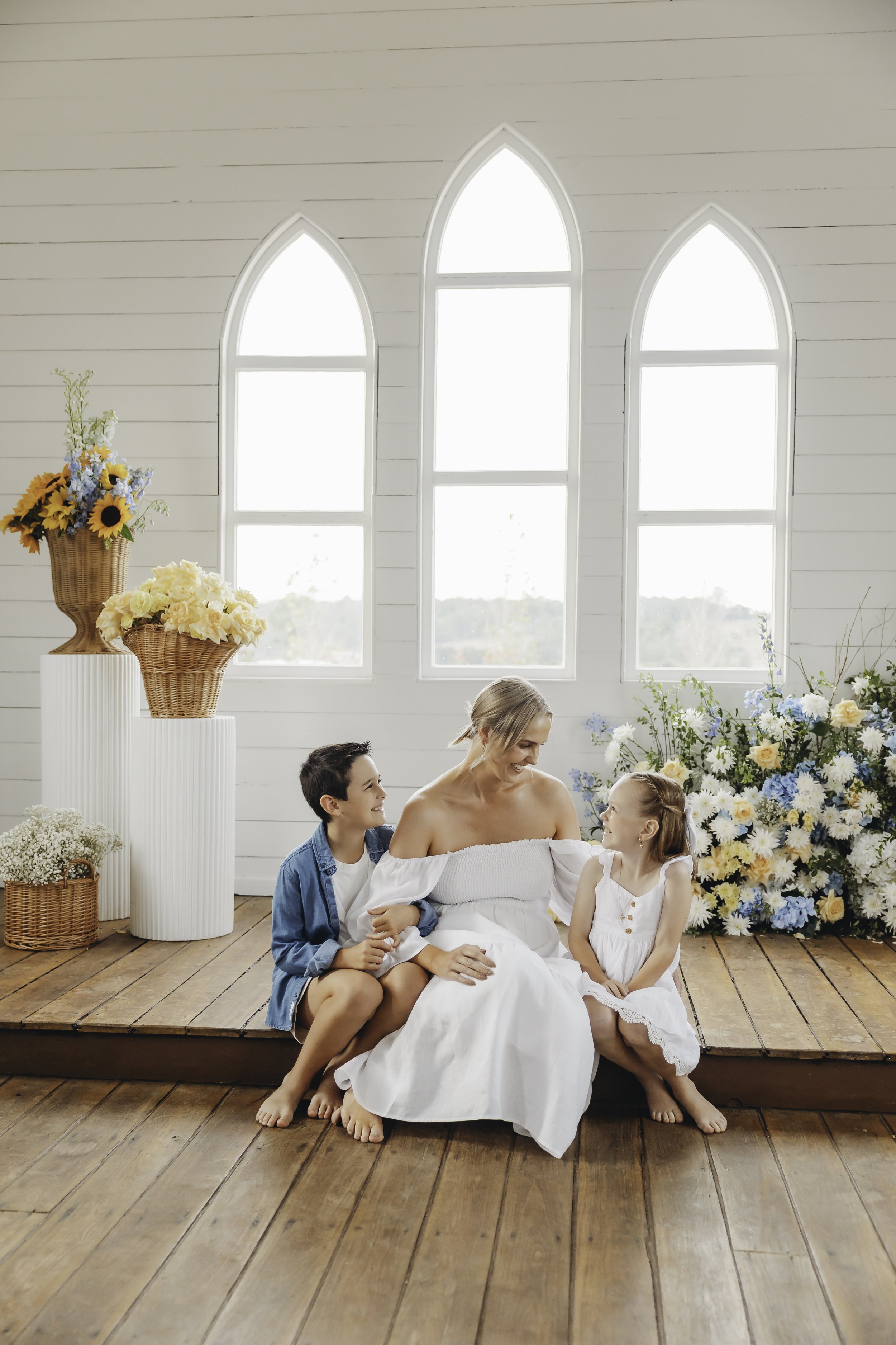 A woman sitting on a wooden stage with two children, a boy and a girl, in a bright, floral room with church-style windows, and floral arrangements.