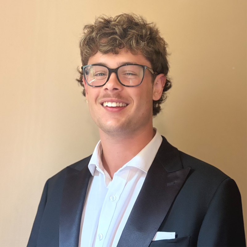 Young man with curly hair and glasses smiling, wearing a tuxedo with a white shirt against a beige background.