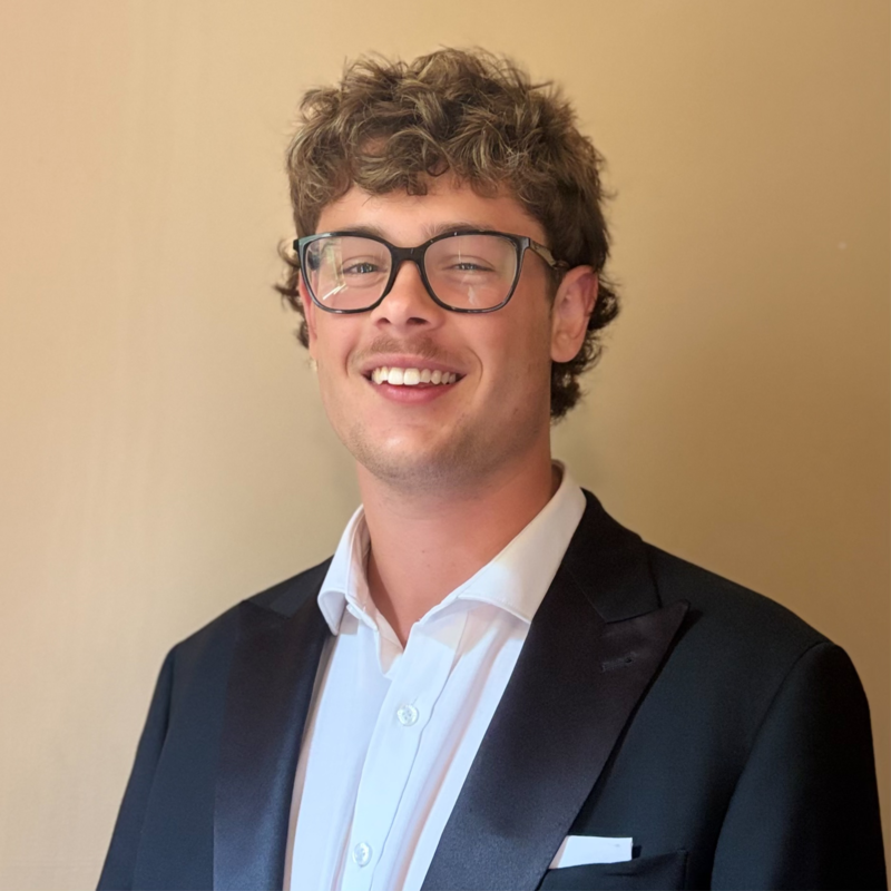 Young man with curly brown hair wearing glasses, a white dress shirt, and a black tuxedo with a satin lapel, smiling at the camera.