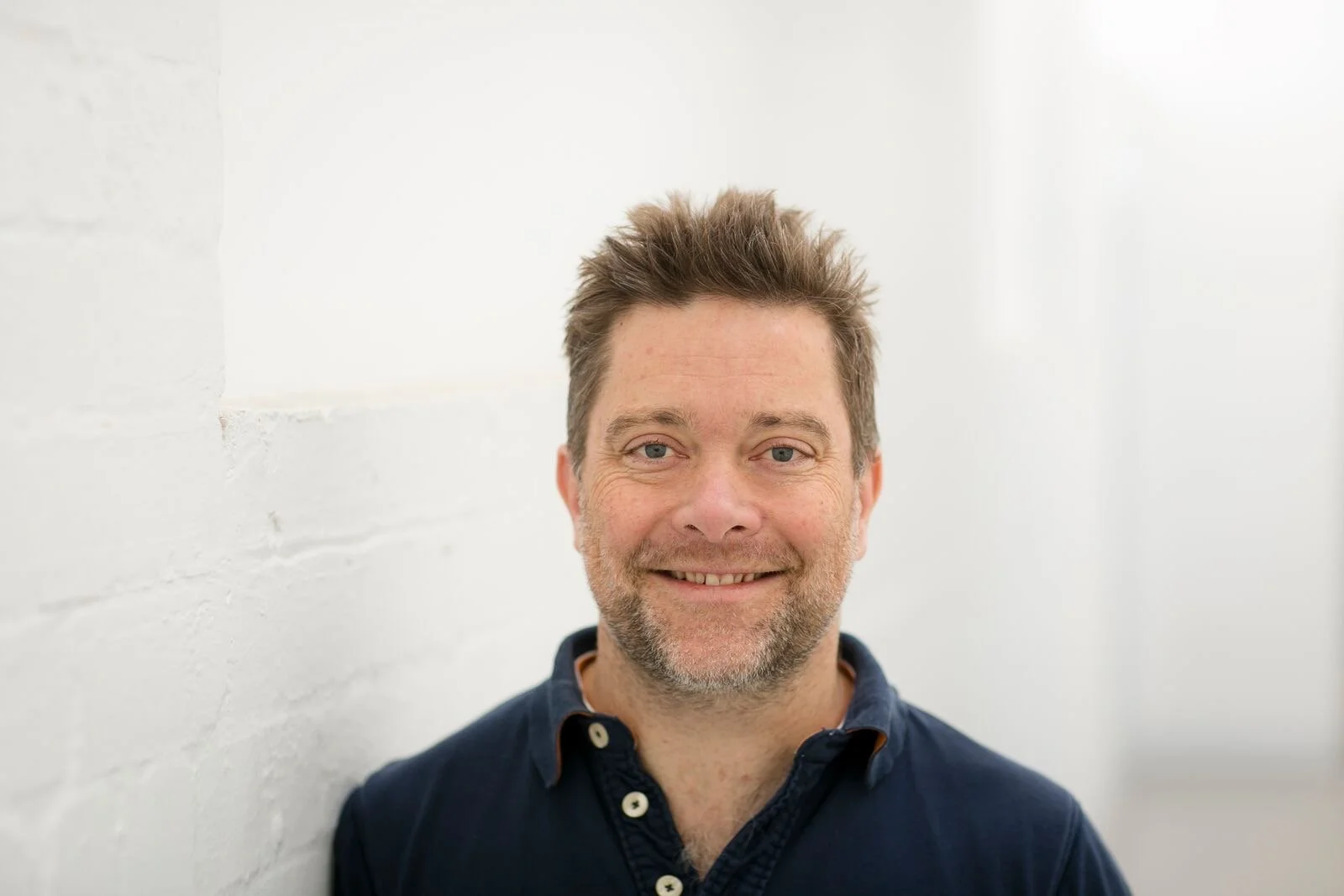 A smiling man with short, spiky brown hair and blue eyes, wearing a navy blue collared shirt, standing in front of a white brick wall.