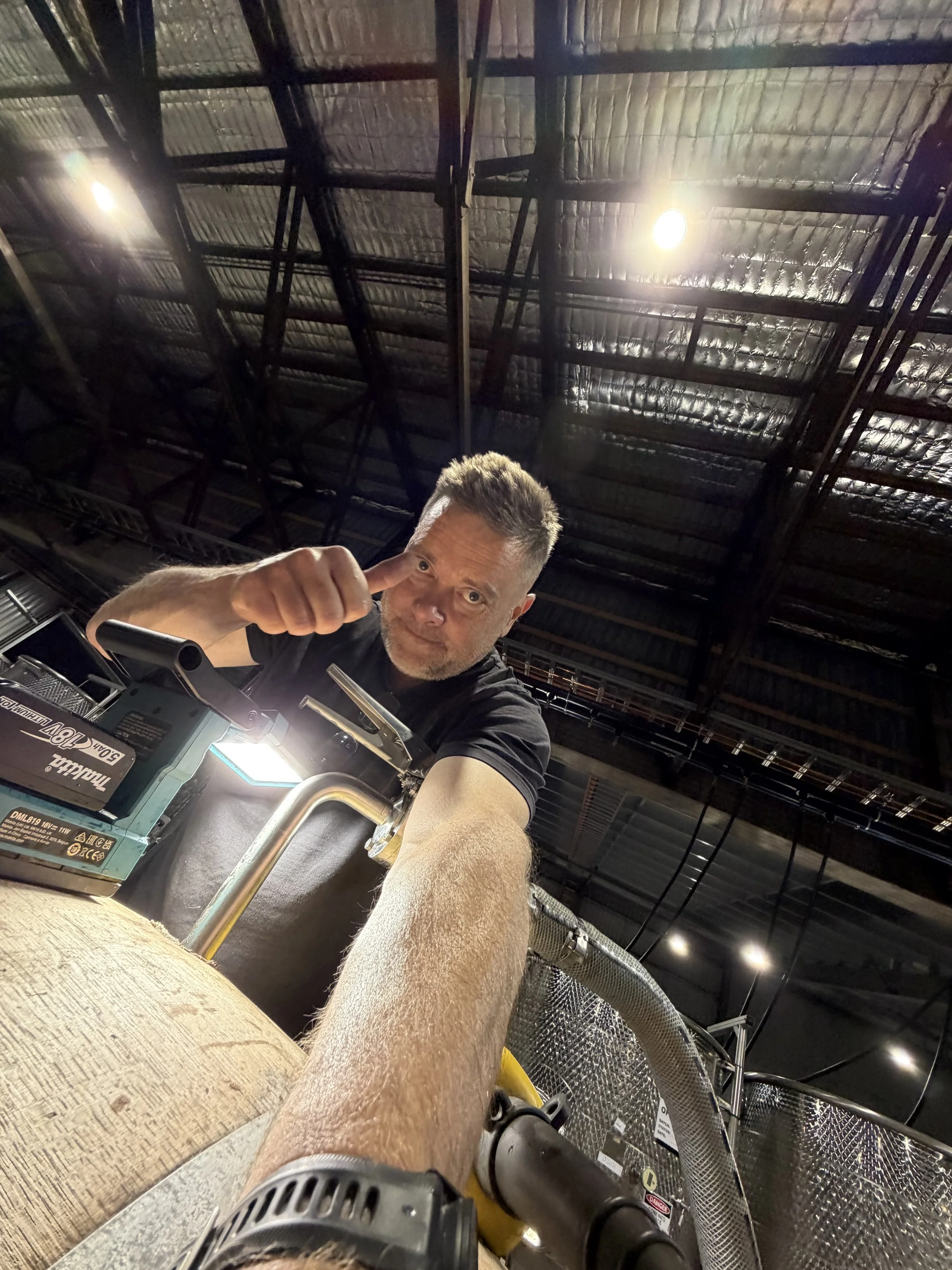 A man with short gray hair and a black shirt sitting at a workbench in an industrial or warehouse setting, giving a thumbs-up. The photo is taken from his perspective, showing his arm, a power tool in front of him, and the high ceiling with metal bea