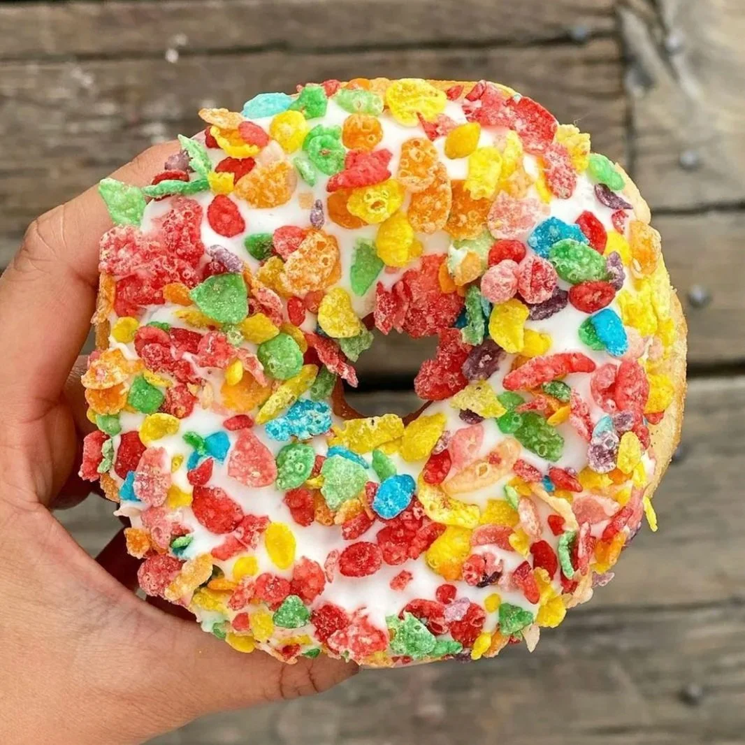 A hand holding a sprinkled donut with white icing and colorful cereal pieces on top, against a wooden background.