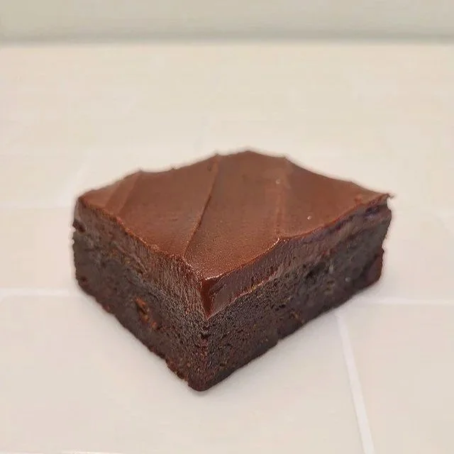 Six pieces of brownie with a glossy chocolate glaze, arranged on a white surface, each piece in a small paper cup.