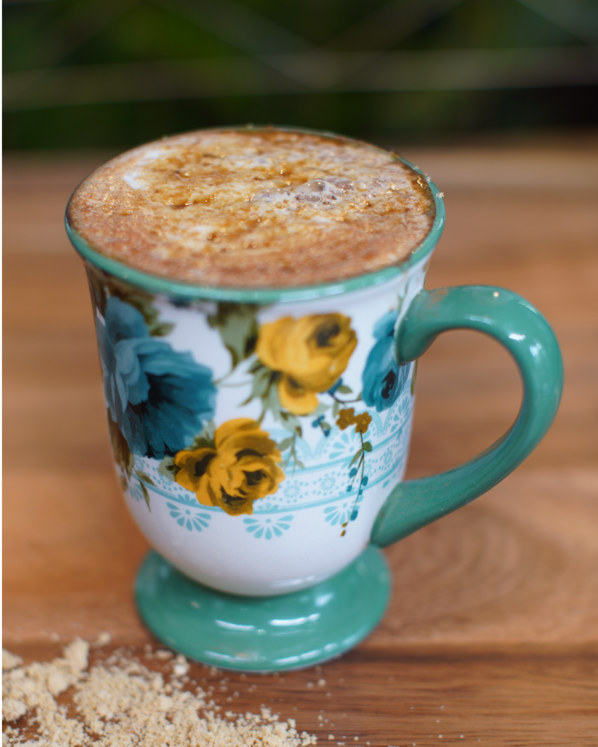 A floral patterned mug filled with a frothy cinnamon latte, placed on a wooden surface with some cinnamon powder scattered nearby.