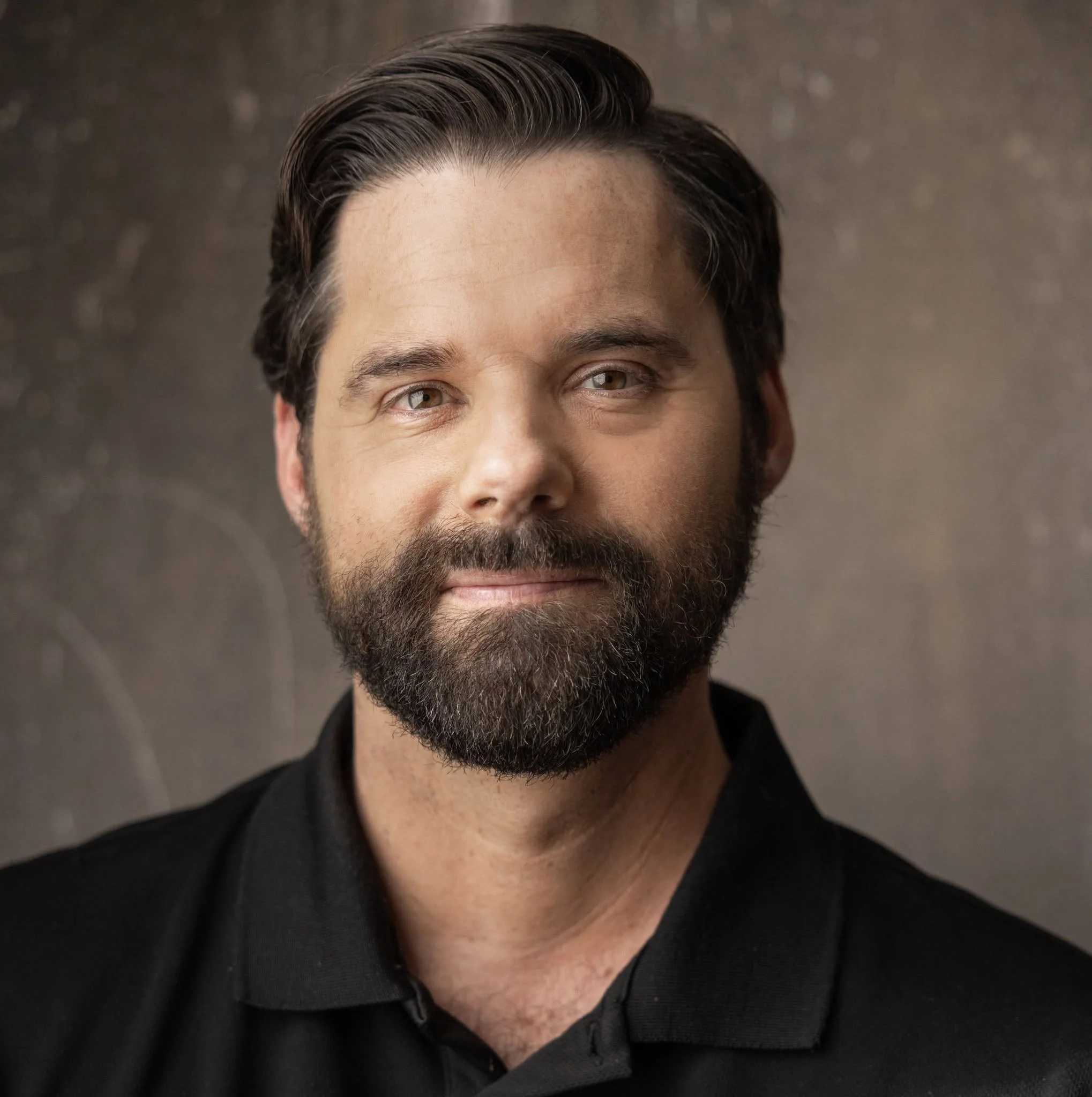 Close-up of a man's face showing brown hair styled back, a beard, and brown eyes, wearing a black shirt.