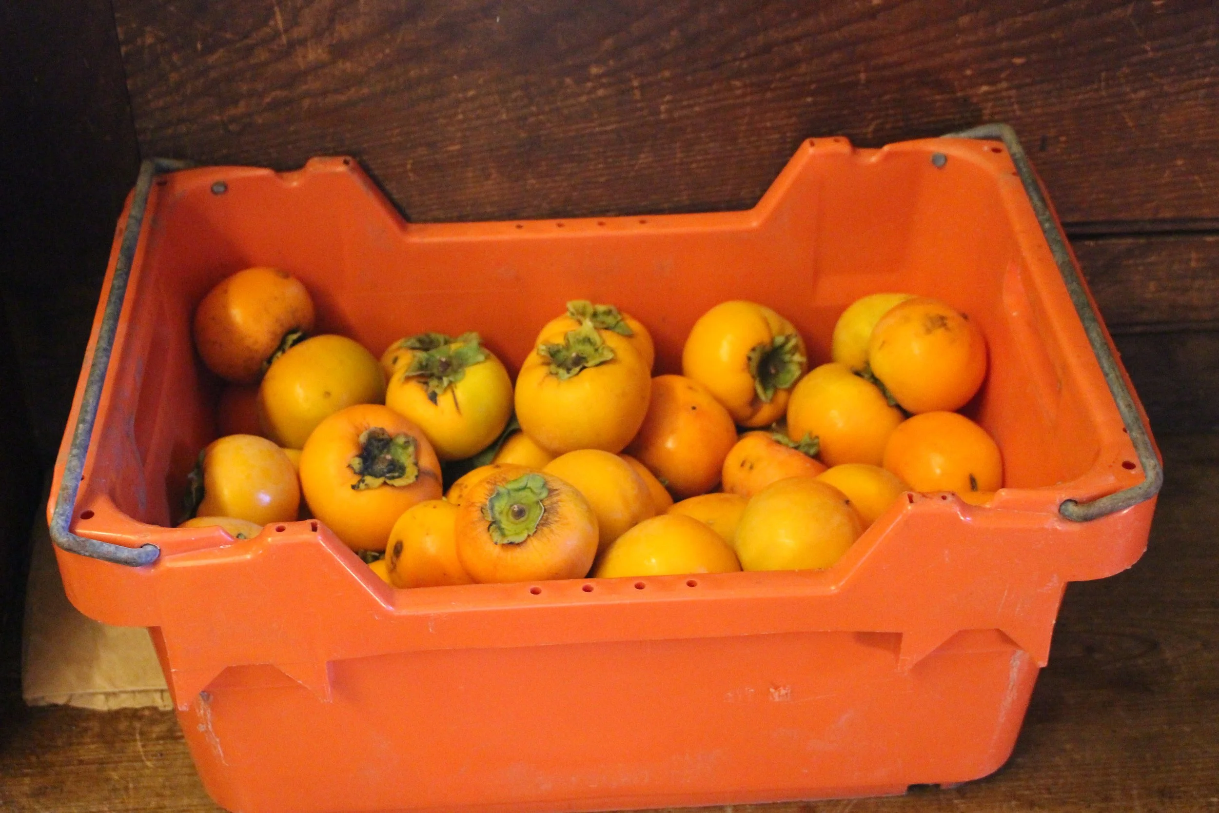 A plastic orange crate filled with yellow persimmons, some with green calyxes, on a dark wooden surface.
