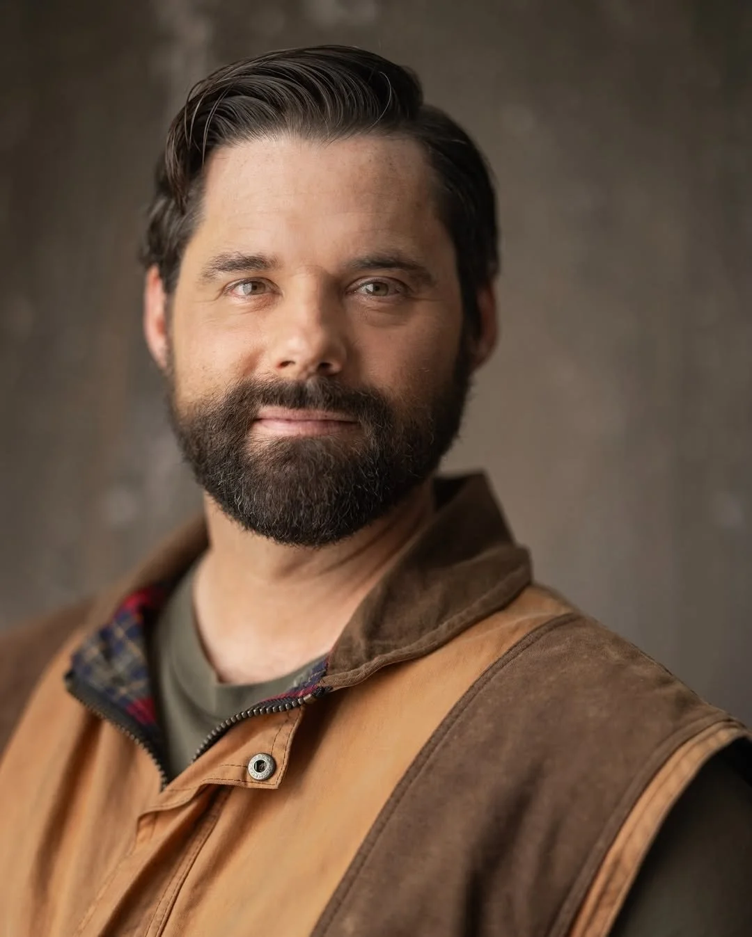 A man with dark hair and a beard, wearing a brown and tan jacket over a green shirt, looking slightly to the side with a neutral expression, against a blurred dark background.