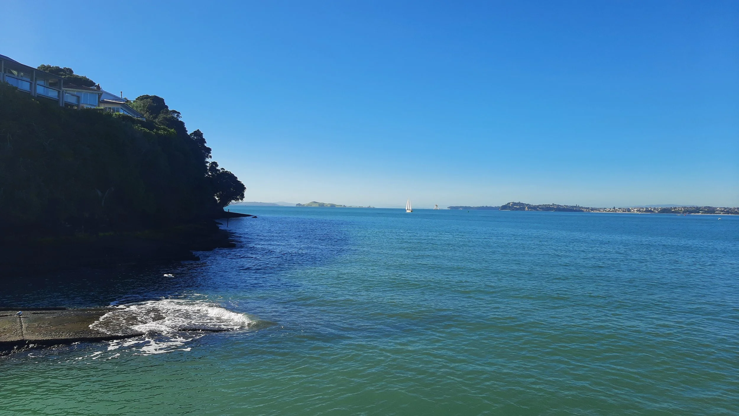A scenic view of a bay with calm blue water, a sailboat in the distance, a green hillside with houses on the left, and distant landforms on the horizon under a clear blue sky.