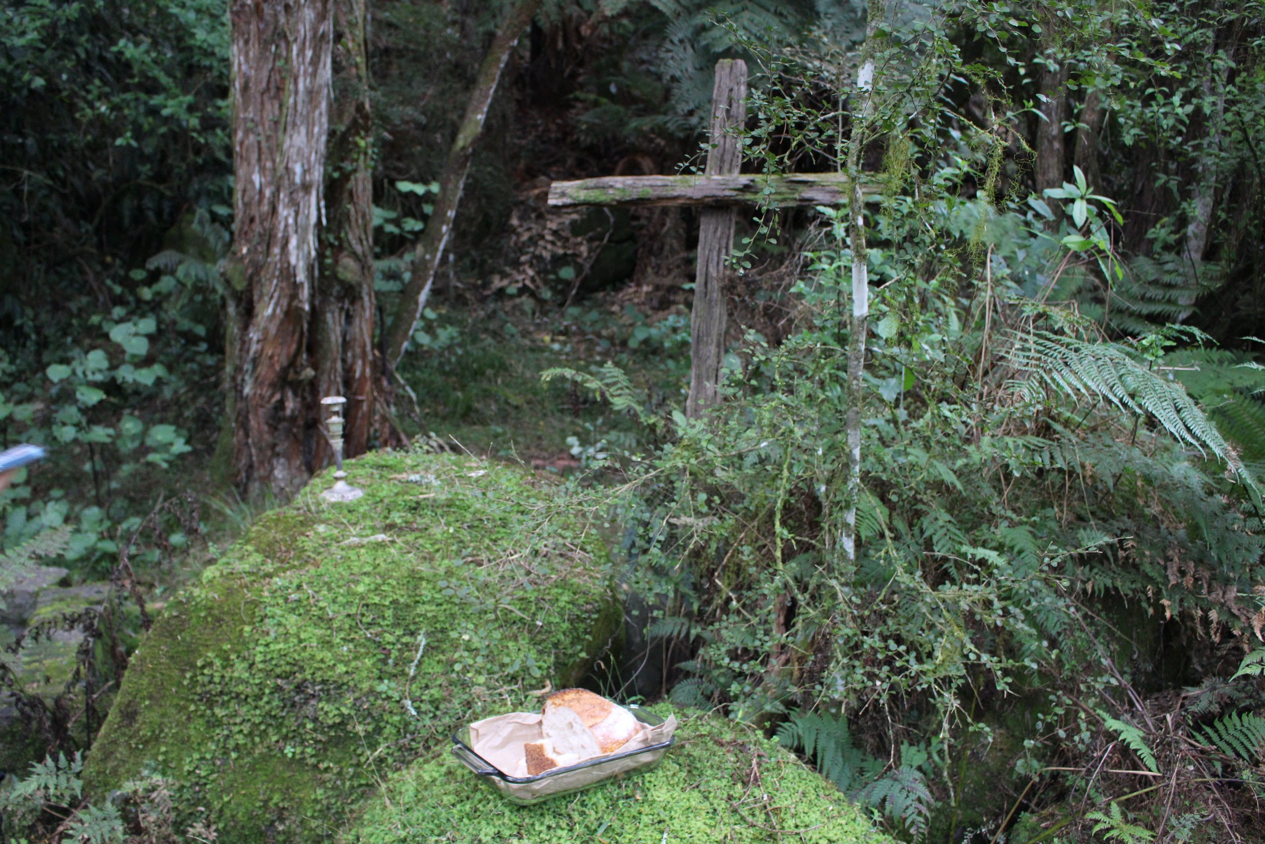 A rustic outdoor table in a lush, green forest setting with moss-covered rocks and leafy plants surrounding it. There is a tray of sliced bread on the ground, and a wooden cross-like structure made from rough logs stands in the background.