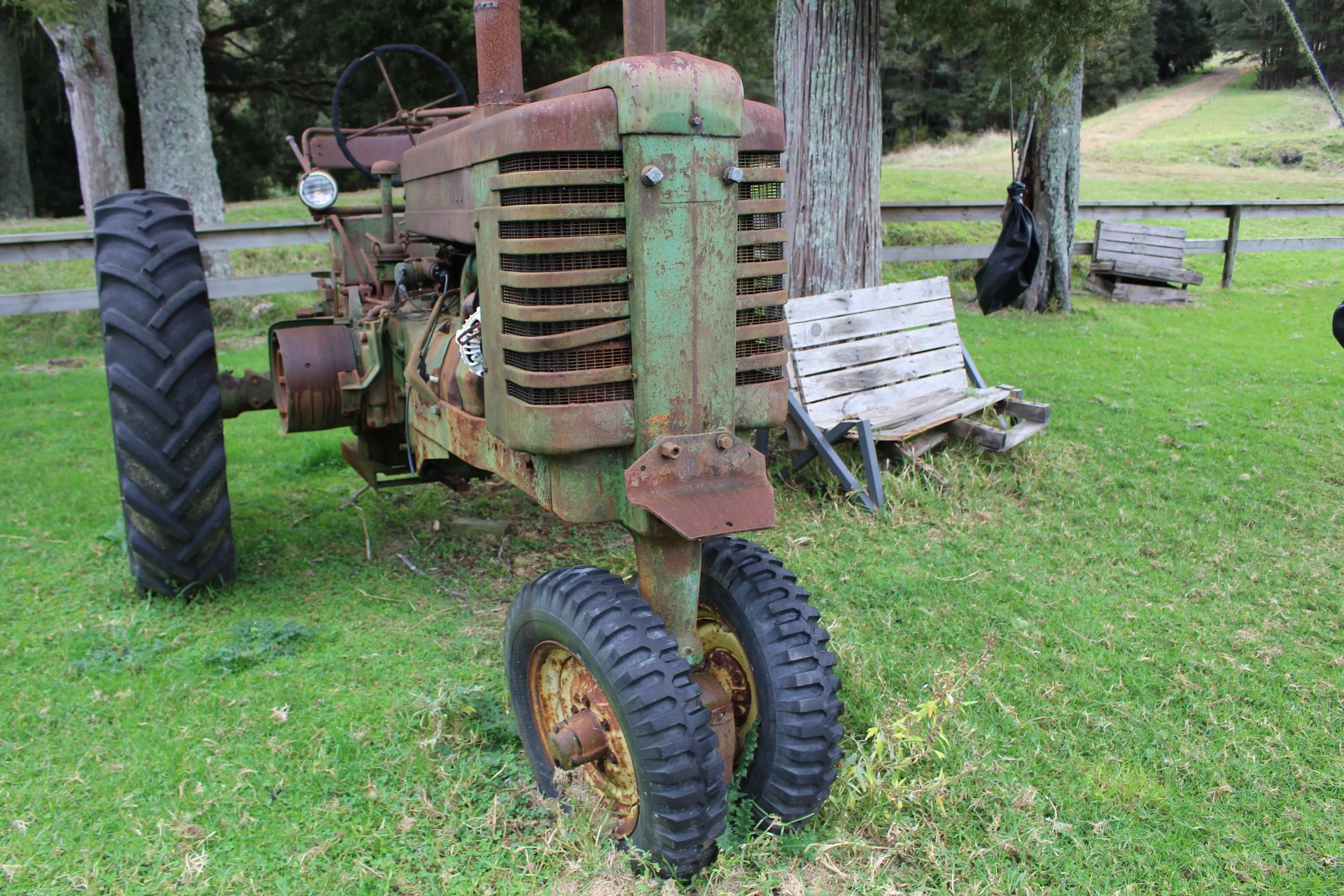An old rusty tractor with large tires and peeling green paint, parked on a grassy area near trees and a wooden bench.