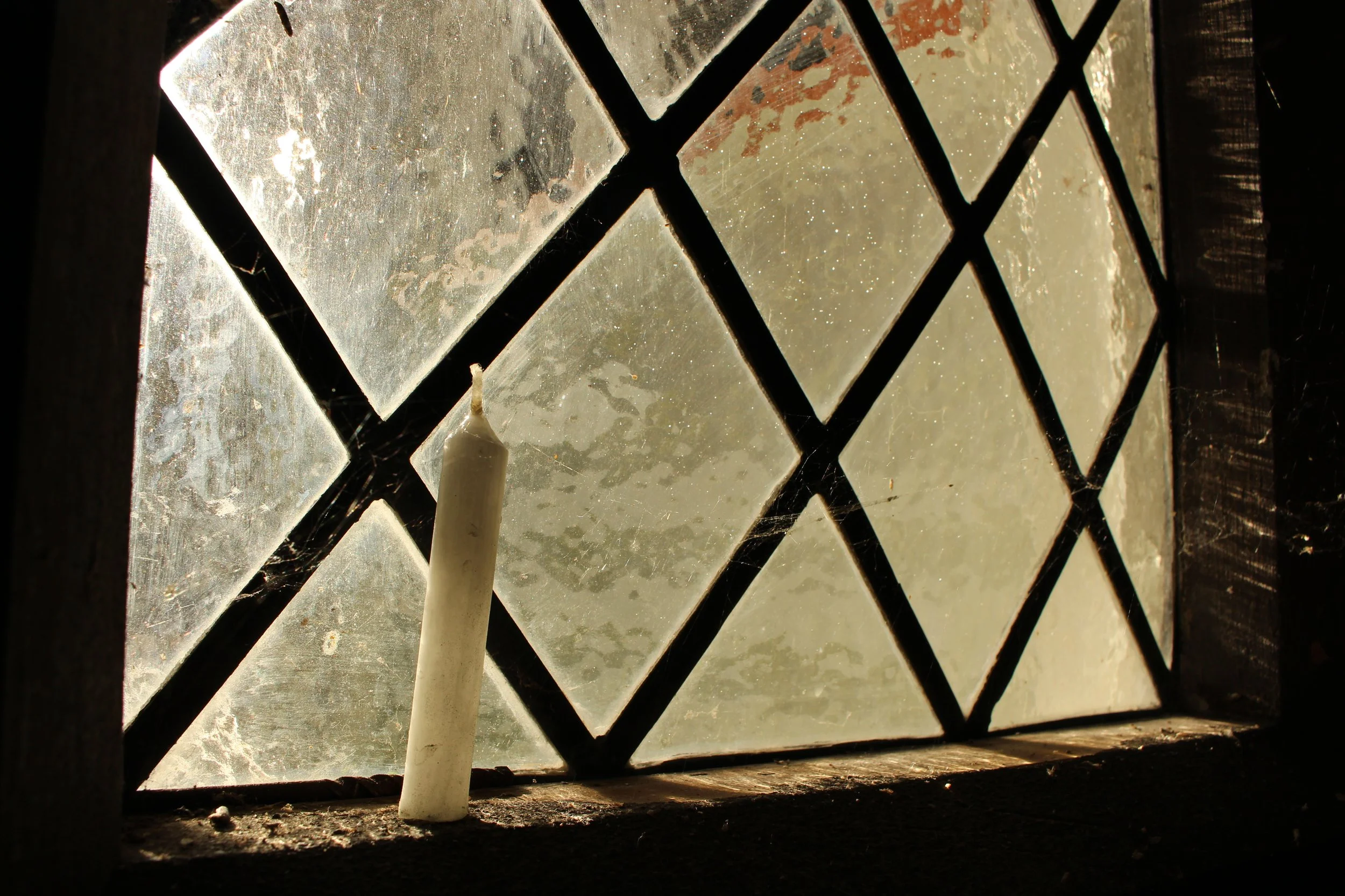 Close-up of dust-covered glass window with black metal framework, illuminated by sunlight, with a white candle placed on the window sill.