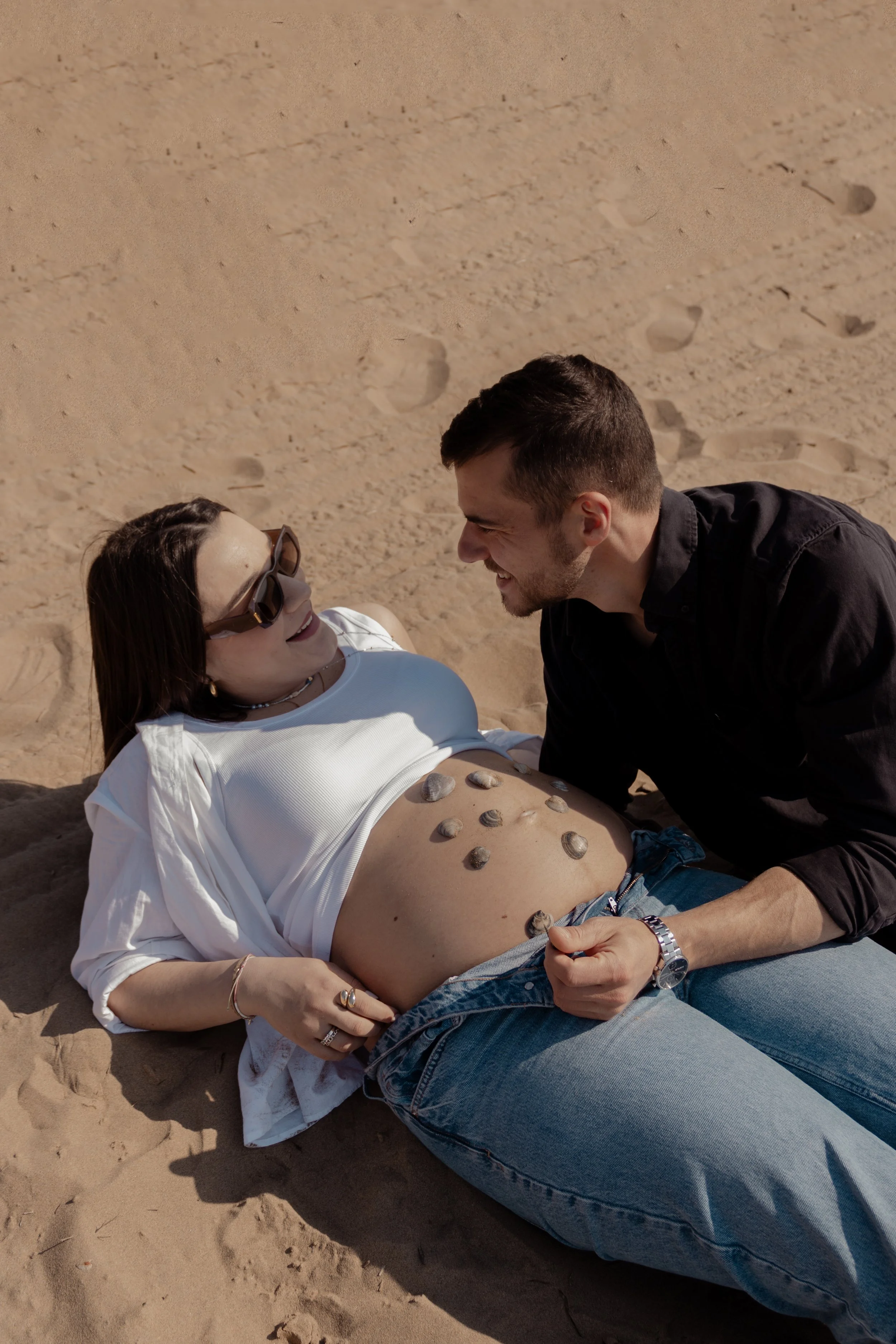 Una donna e un uomo si divertono sulla spiaggia, la donna è sdraiata sulla sabbia con pezzi di conchiglie sul ventre, entrambi sorridono e si guardano felici.