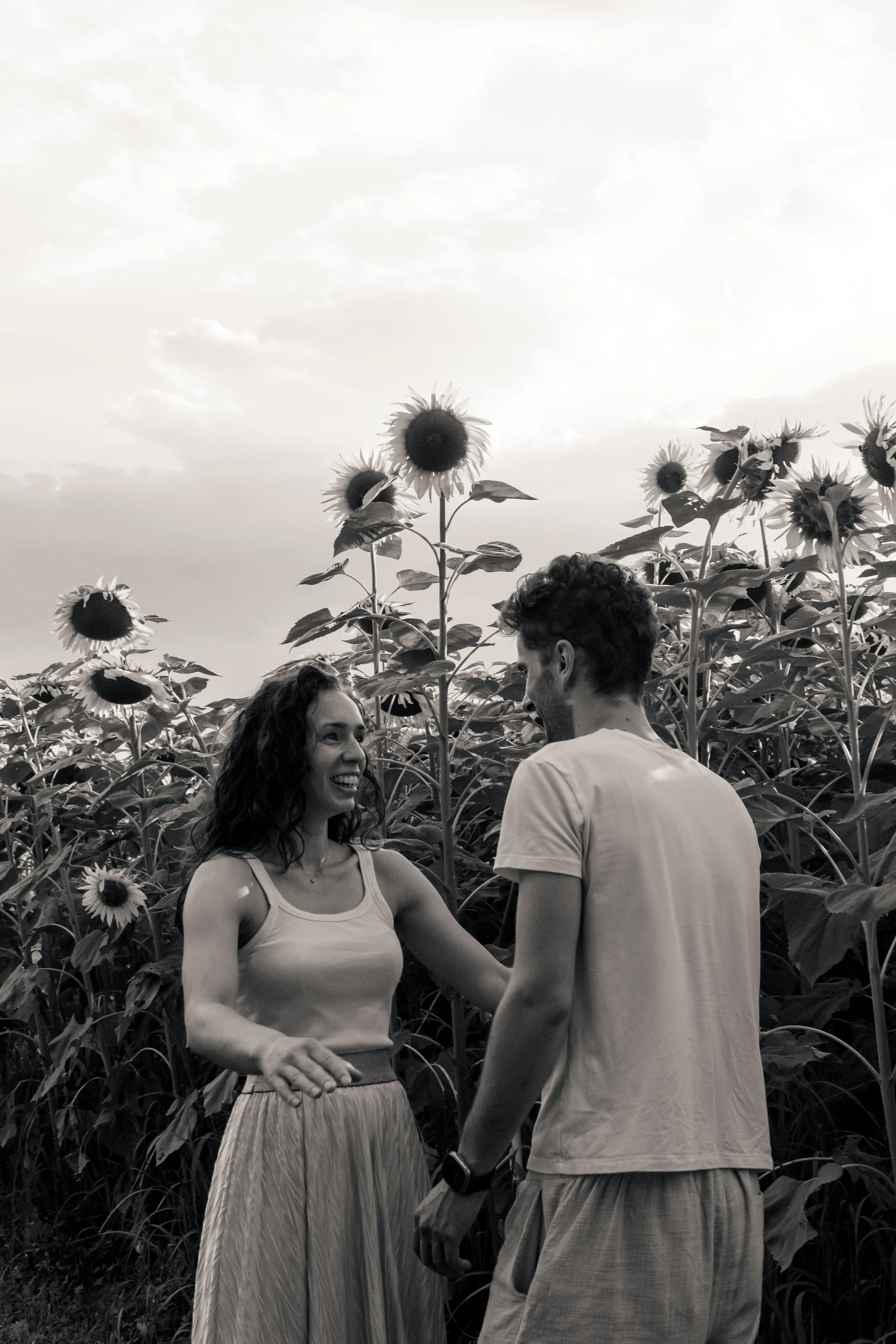 Due persone, una donna e un uomo, stanno sorridendo e si tengono per le mani in un campo di girasoli, con il cielo nuvoloso sullo sfondo.