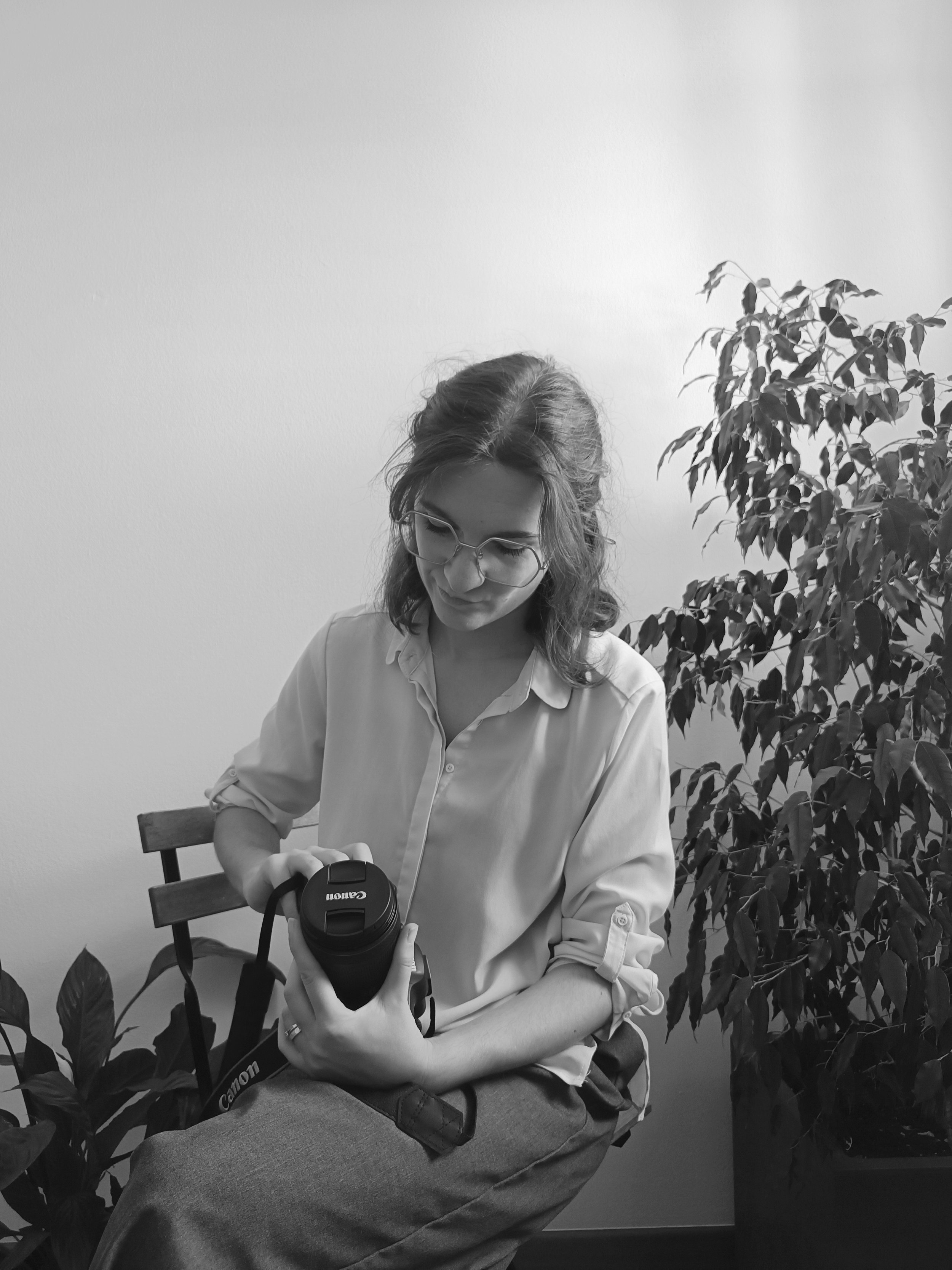 Una donna con occhiali e camicia bianca seduta su una sedia, tiene in mano una fotocamera Canon e sembra prepararsi a scattare una foto. Sullo sfondo, una pianta verde e una parete bianca.