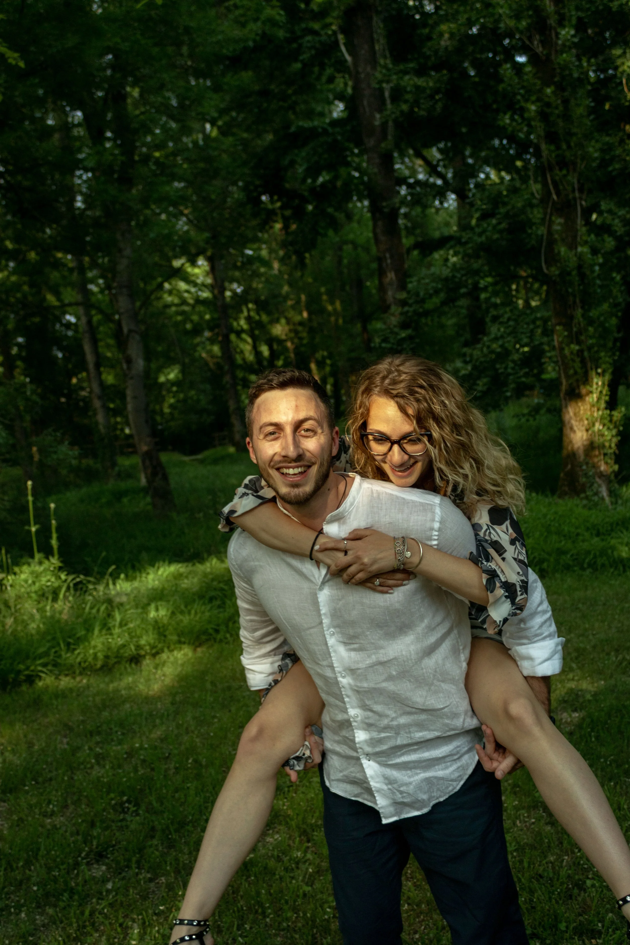Una coppia di giovani si diverte in un parco con alberi verdi, con il ragazzo che sorride e la ragazza con gli occhiali che lo abbraccia da dietro, mentre un'altra persona sta vicino a loro.