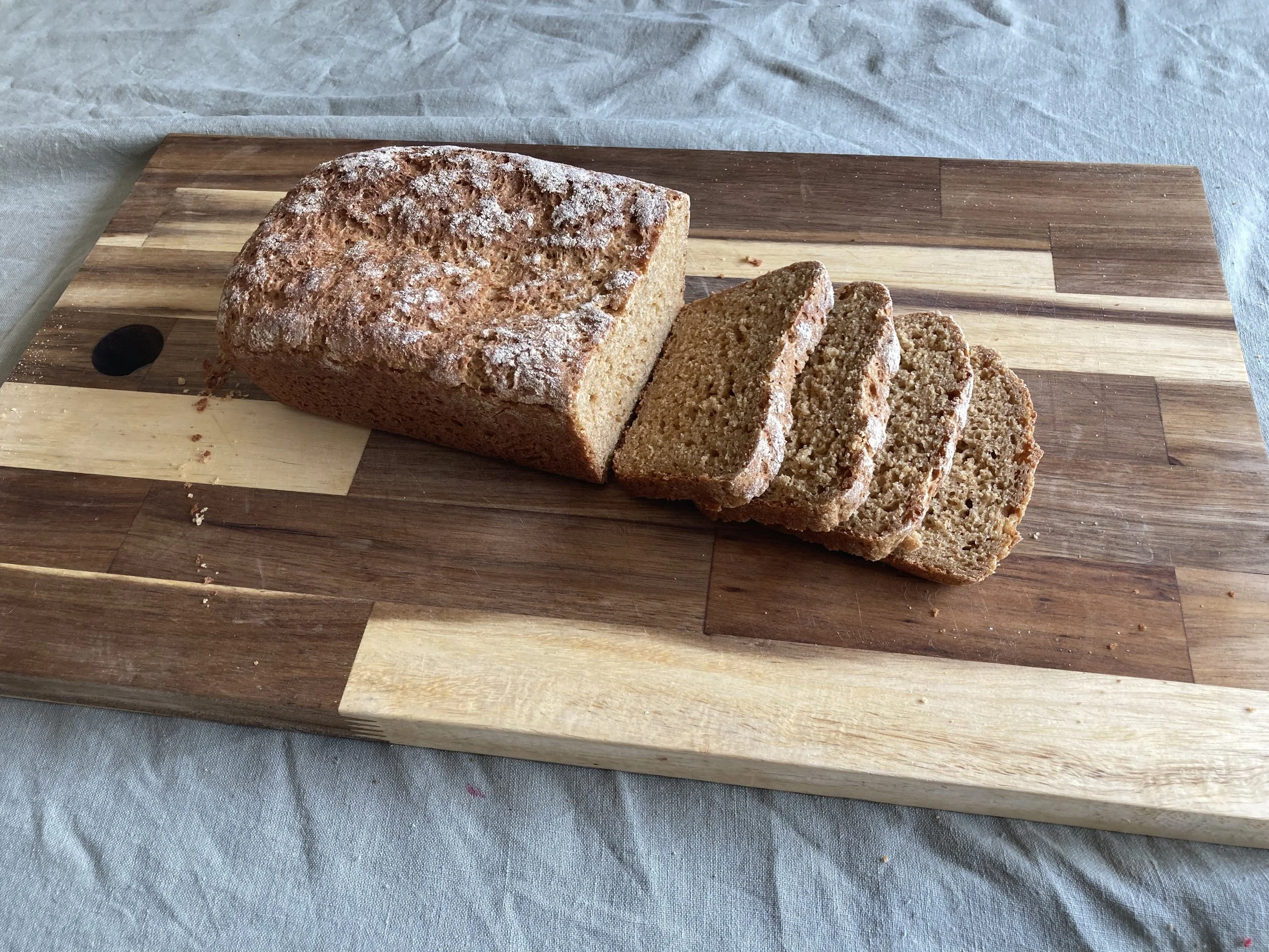 A loaf of organic, fresh-milled einkorn bread, resting on a wooden cutting board with three slices cut from it.