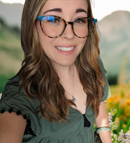 Young woman with long brown hair, glasses, and a green top smiling outdoors with hills and flowers in the background.