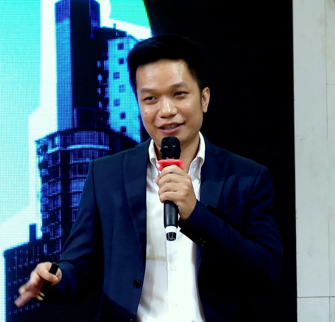A young man in a suit speaking into a microphone during a presentation or speech, with a colorful digital display of a building in the background.