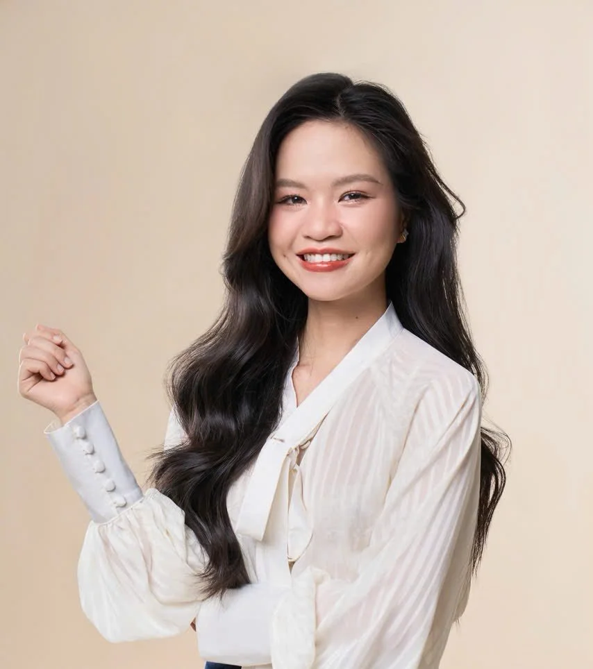 A Vietnamese woman with long, wavy black hair, smiling, wearing a cream-colored blouse with a bow tie, standing against a neutral beige background.