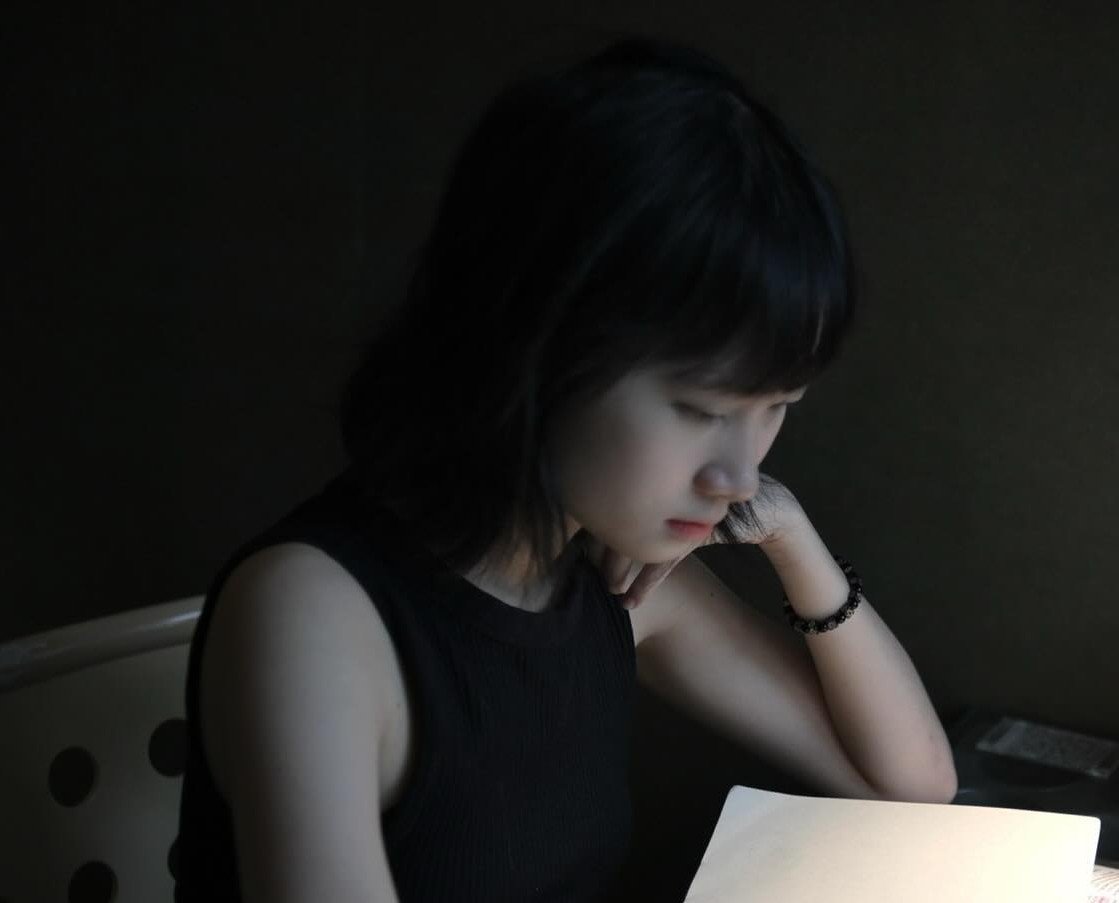 A young woman with shoulder-length dark hair and a black sleeveless top is sitting at a table, looking down at a notebook or paper, with her head resting on her left hand.