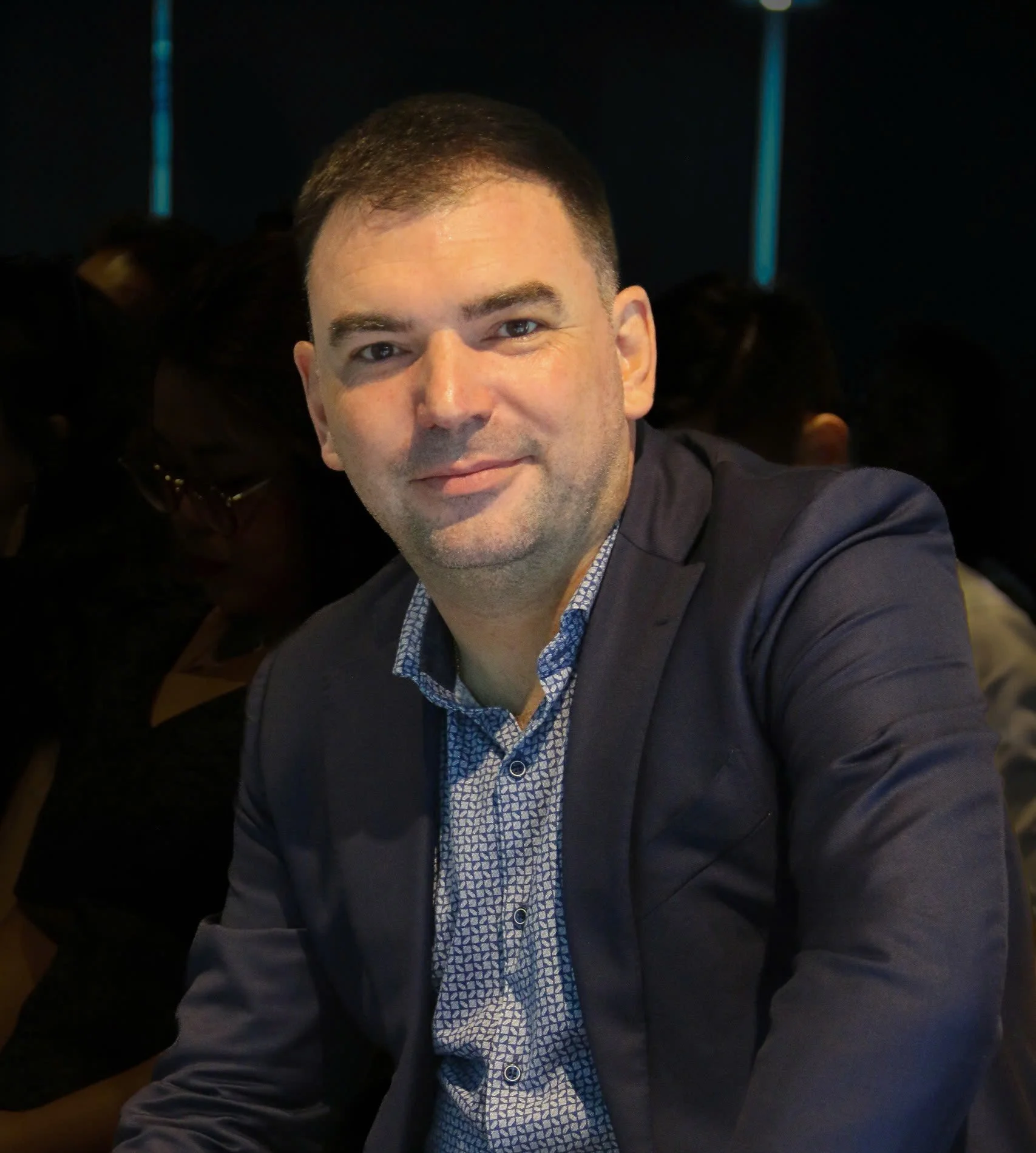 A man with short dark hair and a trimmed beard, wearing a suit jacket and a patterned dress shirt, smiling at the camera in a dark indoor setting.