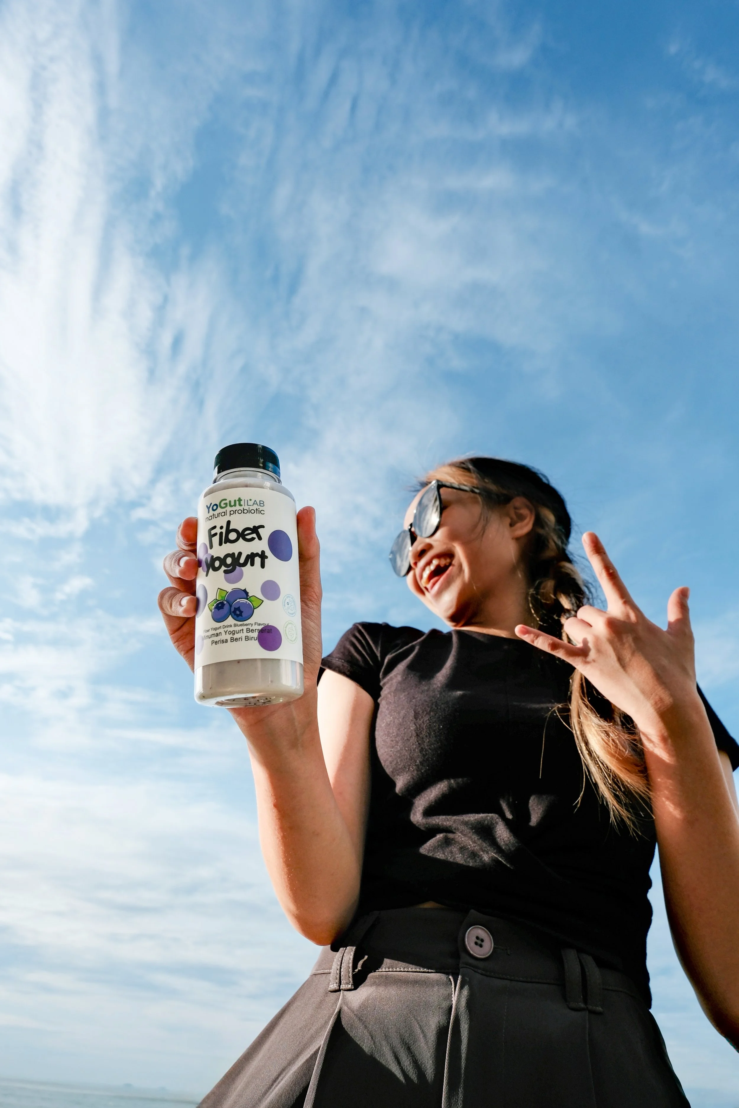 A woman smiling and wearing sunglasses, holds a bottle of YoGutLab's yogurt with a blue sky and clouds in the background.