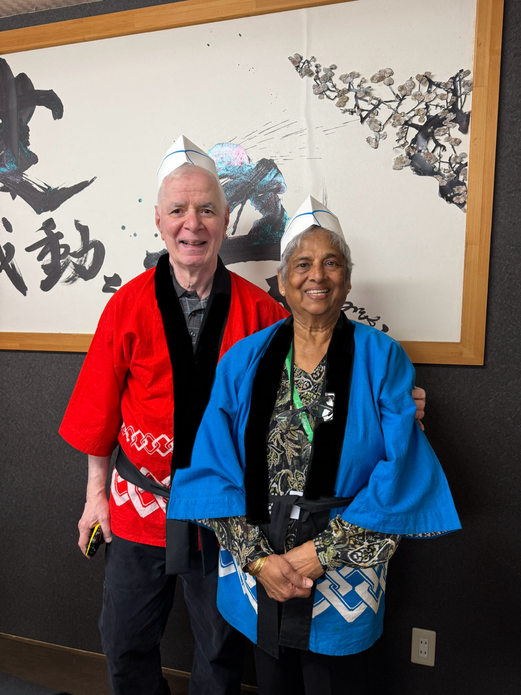 Two older adults, a man and a woman, standing close together, both wearing traditional Japanese attire and white paper hats. They are smiling in front of a large Japanese-style calligraphy and artwork on the wall.