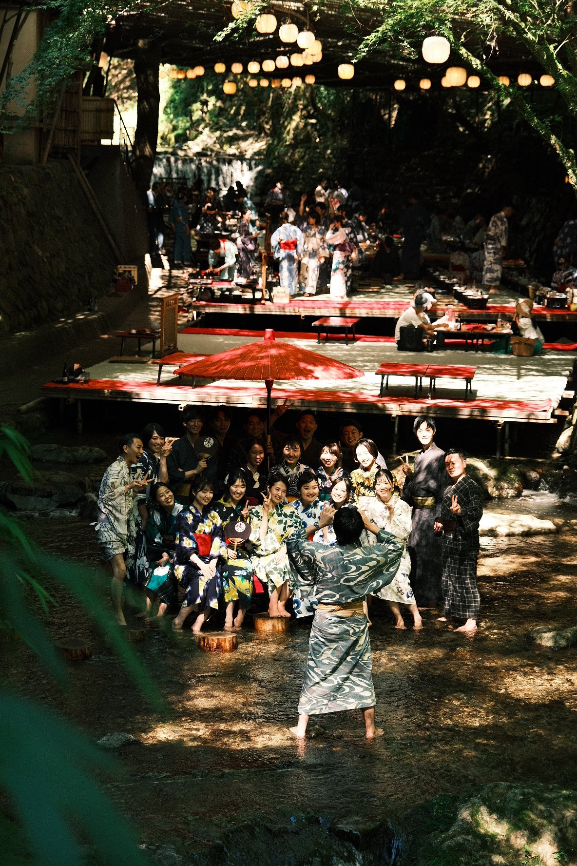 Group of people in traditional Japanese yukatas enjoying a summer festival by a river. Some are standing in the water, while others are sitting or standing near red and black tables set up along the riverbank. Lanterns hang overhead, creating a festive atmosphere.