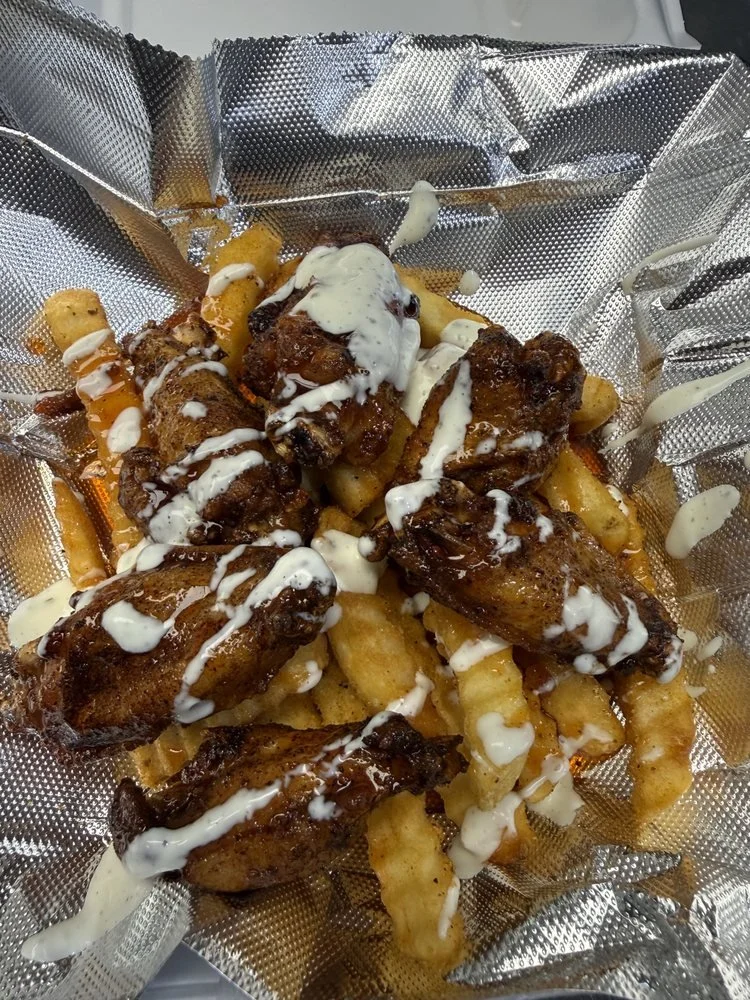 Basket with fried chicken wings, crinkle-cut fries, topped with creamy white sauce in a foil-lined container.