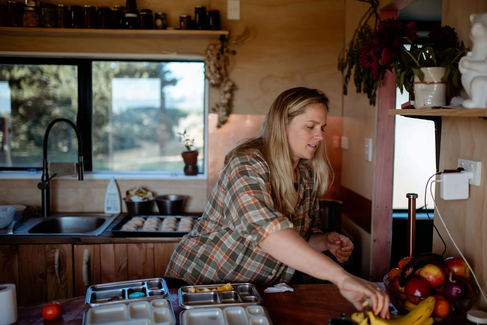 A woman with long blonde hair wearing a plaid shirt is preparing food in a kitchen. There are apples and bananas on the counter, and a window behind her showing trees outside.