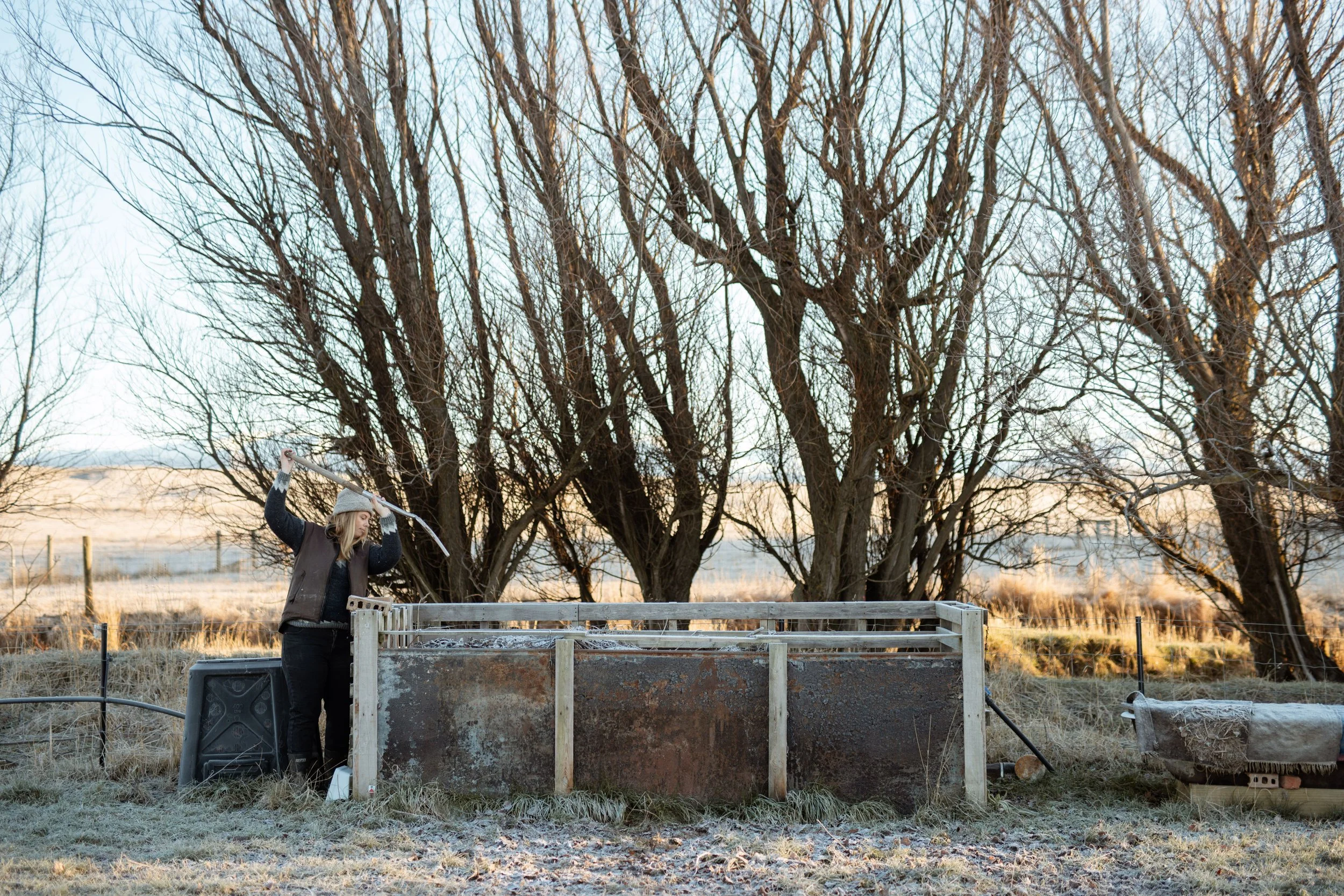 A woman in a jacket and hat is using gardening tools to work with a compost bin outdoors, near leafless trees at sunset.