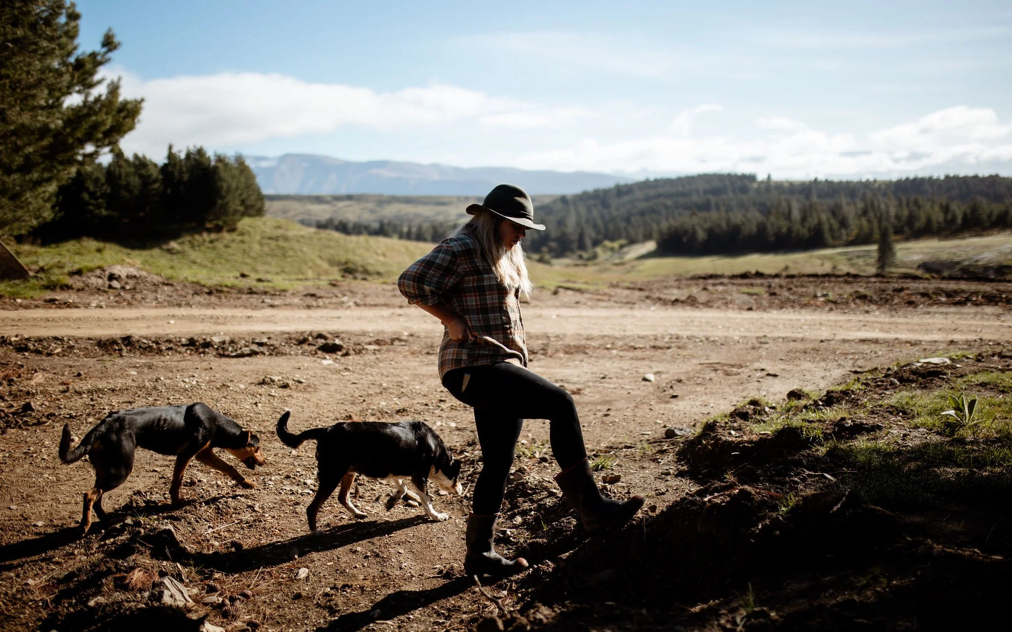 A woman kneels on the ground with her two dogs in a rural outdoor setting, surrounded by trees and mountains in the background.