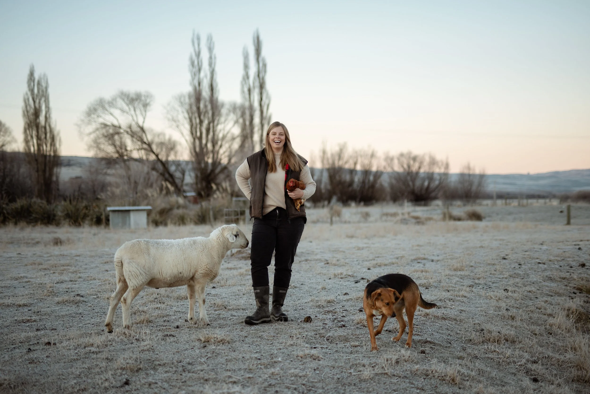 A woman laughing and walking with three dogs in a rural, frosty field during daytime, with trees and hills in the background.