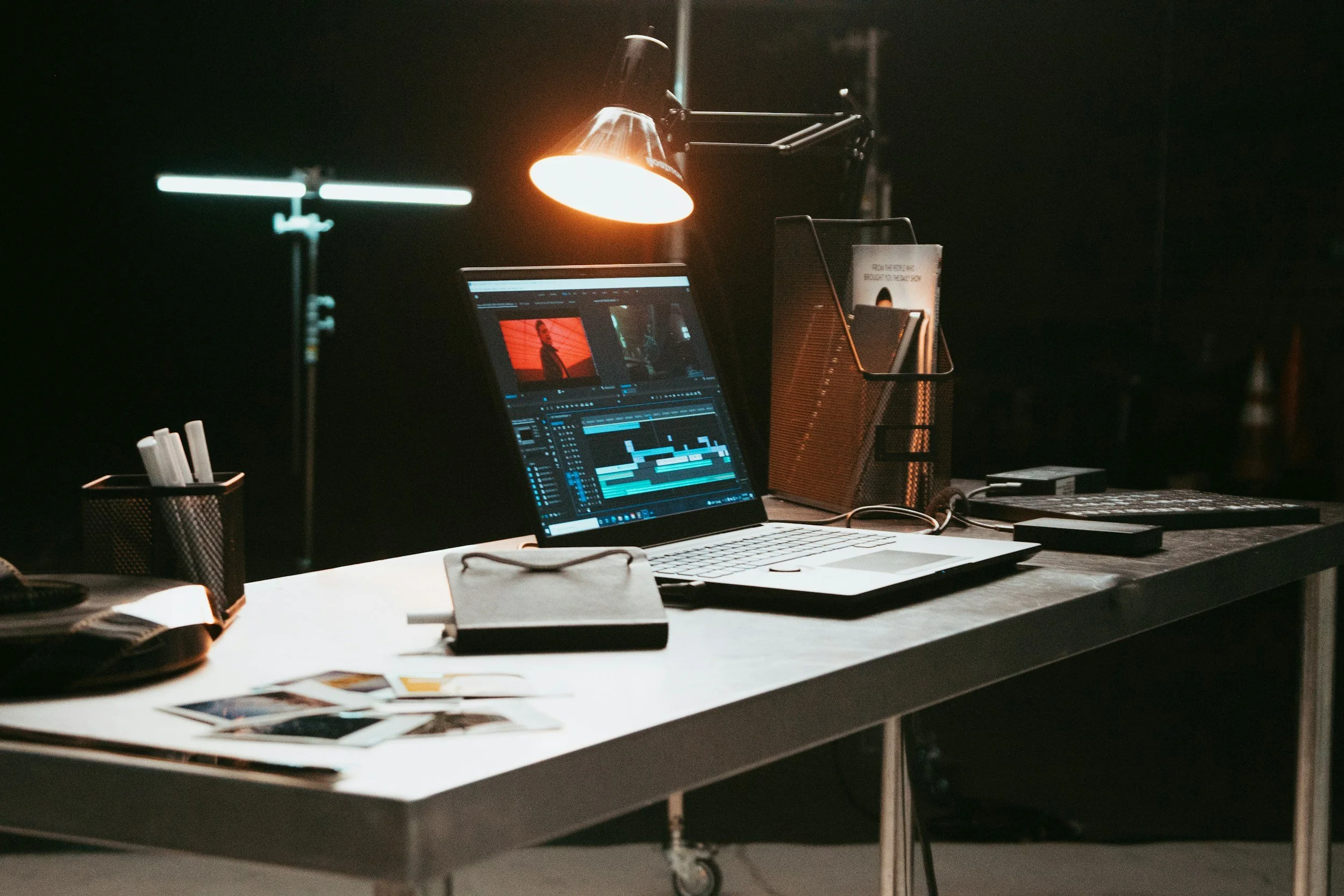 A dimly lit work space with a laptop displaying video editing software, a desktop monitor, and various office supplies on a cluttered desk.
