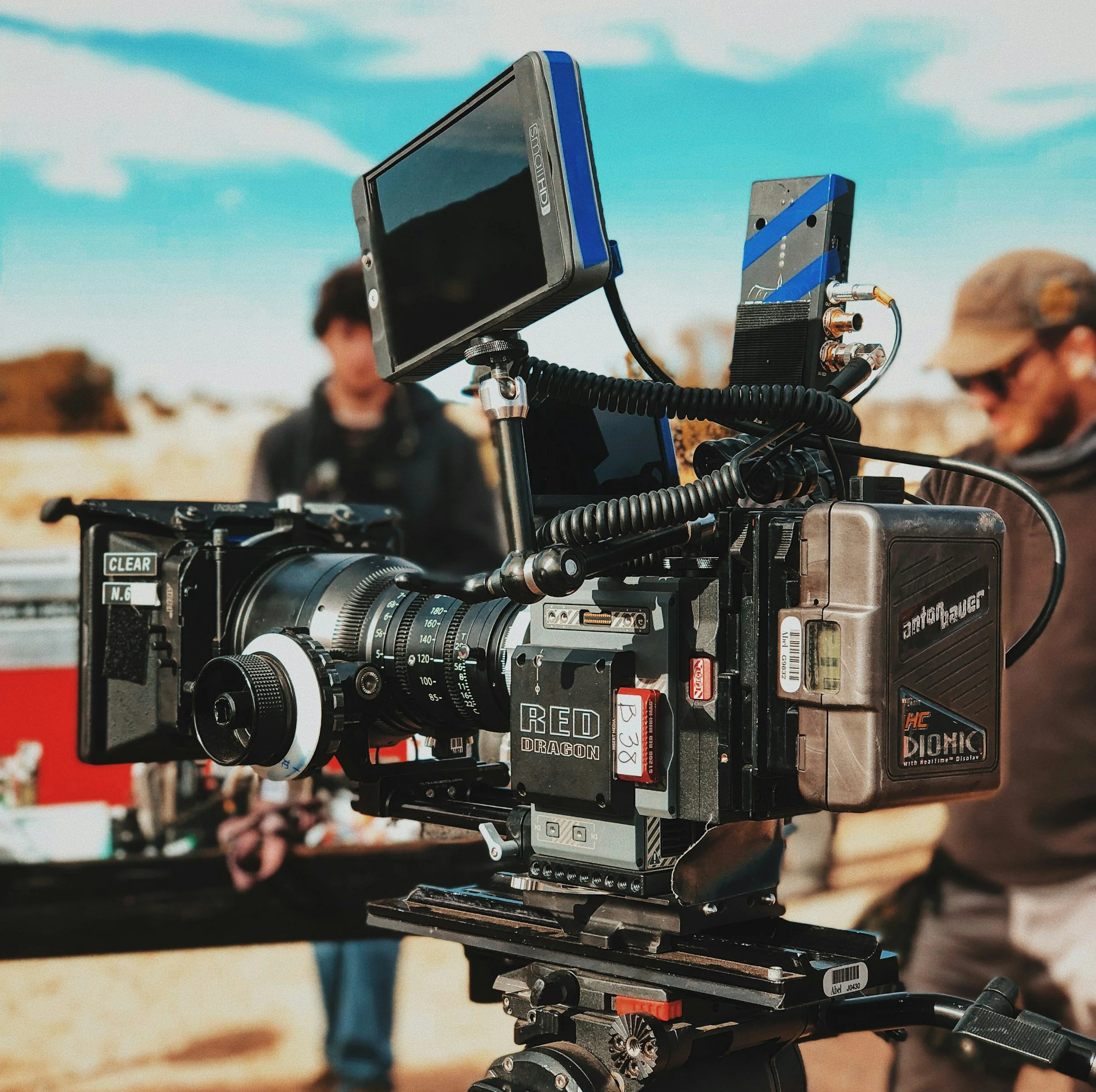 Close-up of a professional film camera film set, with a matte box, lens, and monitor, on a set with people and outdoor background.