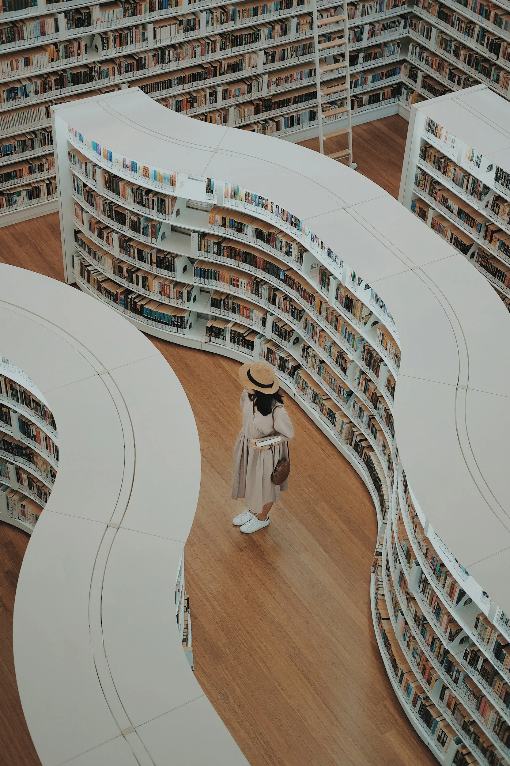 A person with long dark hair wearing a wide-brimmed hat, long beige dress, white sneakers, and carrying a brown bag is browsing books on a curved white bookshelf in a library. The library has wooden floors and multiple white bookshelves filled with books.
