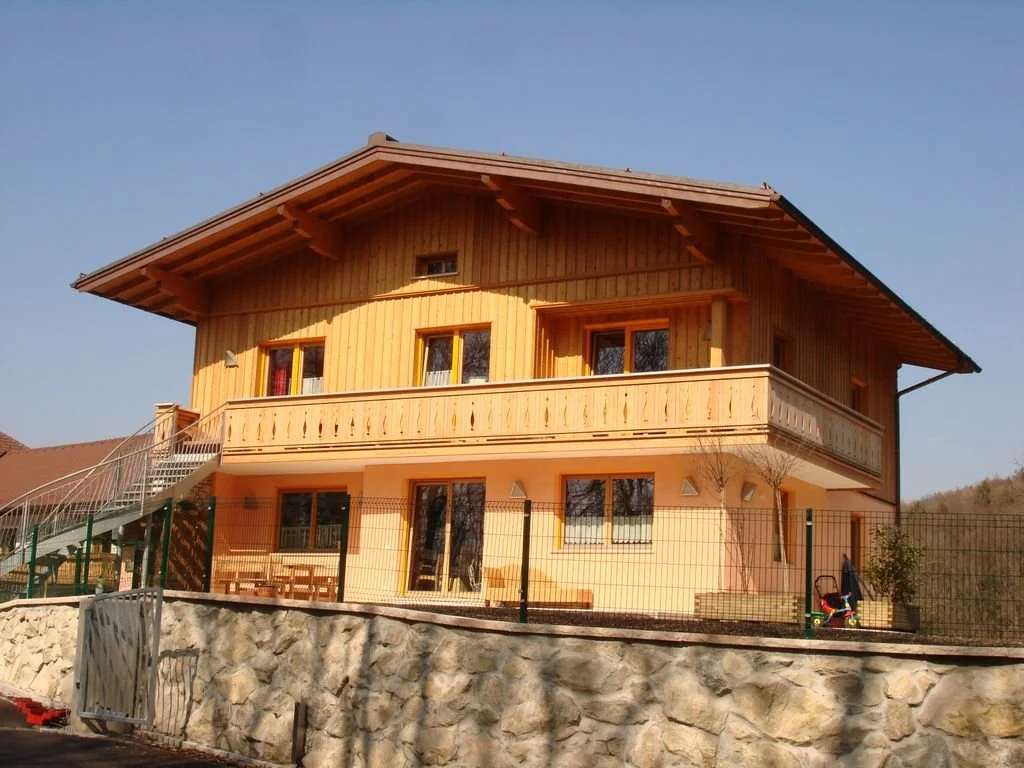 Holzhaus mit Balkon und steiniger Grundmauer, rund um ein Gitterzaun.