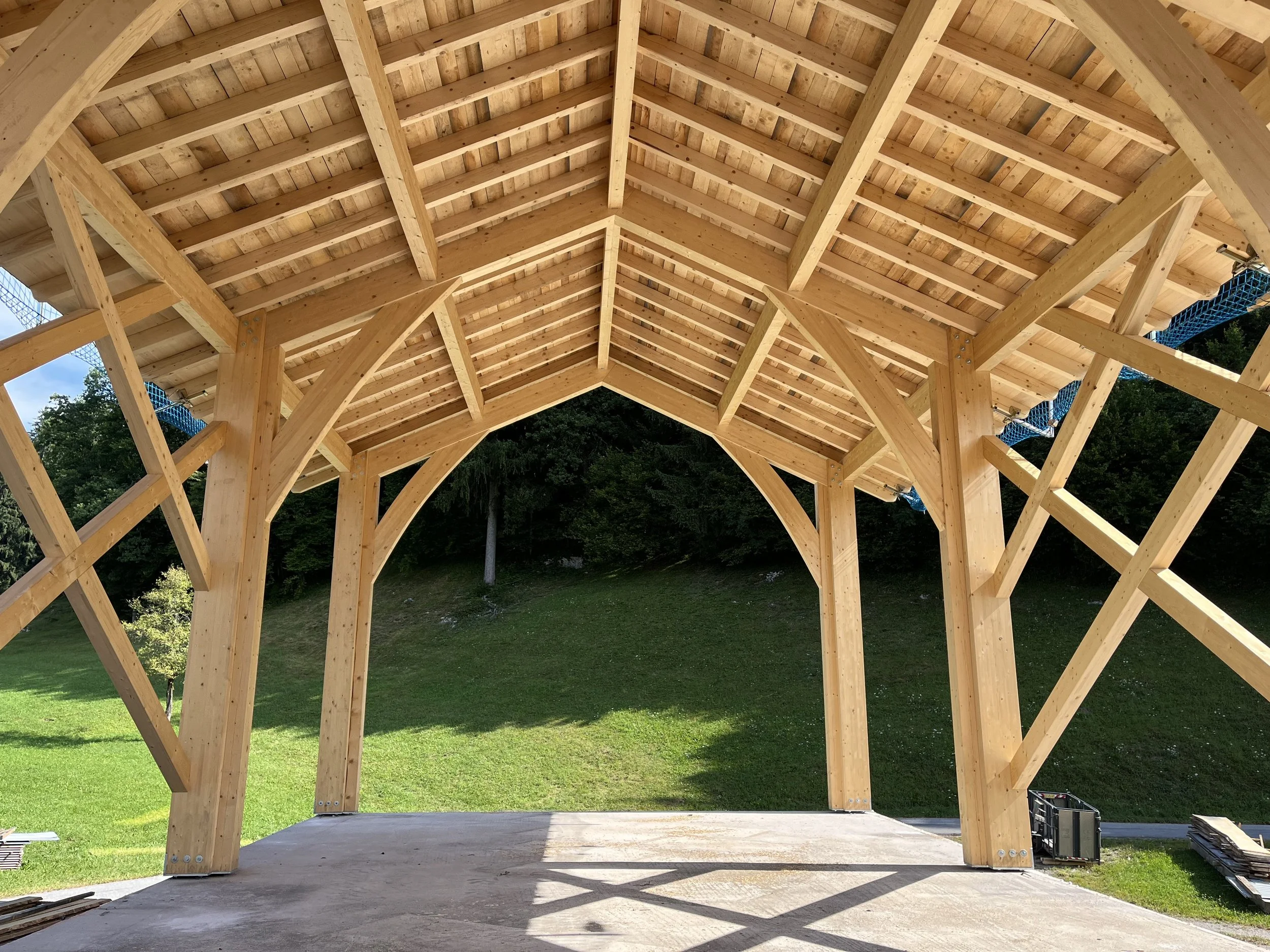 Holzunterstand mit gekreuzten Holzstützen und einem offenen Eingang, im Freien auf einer grünen Wiese, bei sonnigem Wetter.