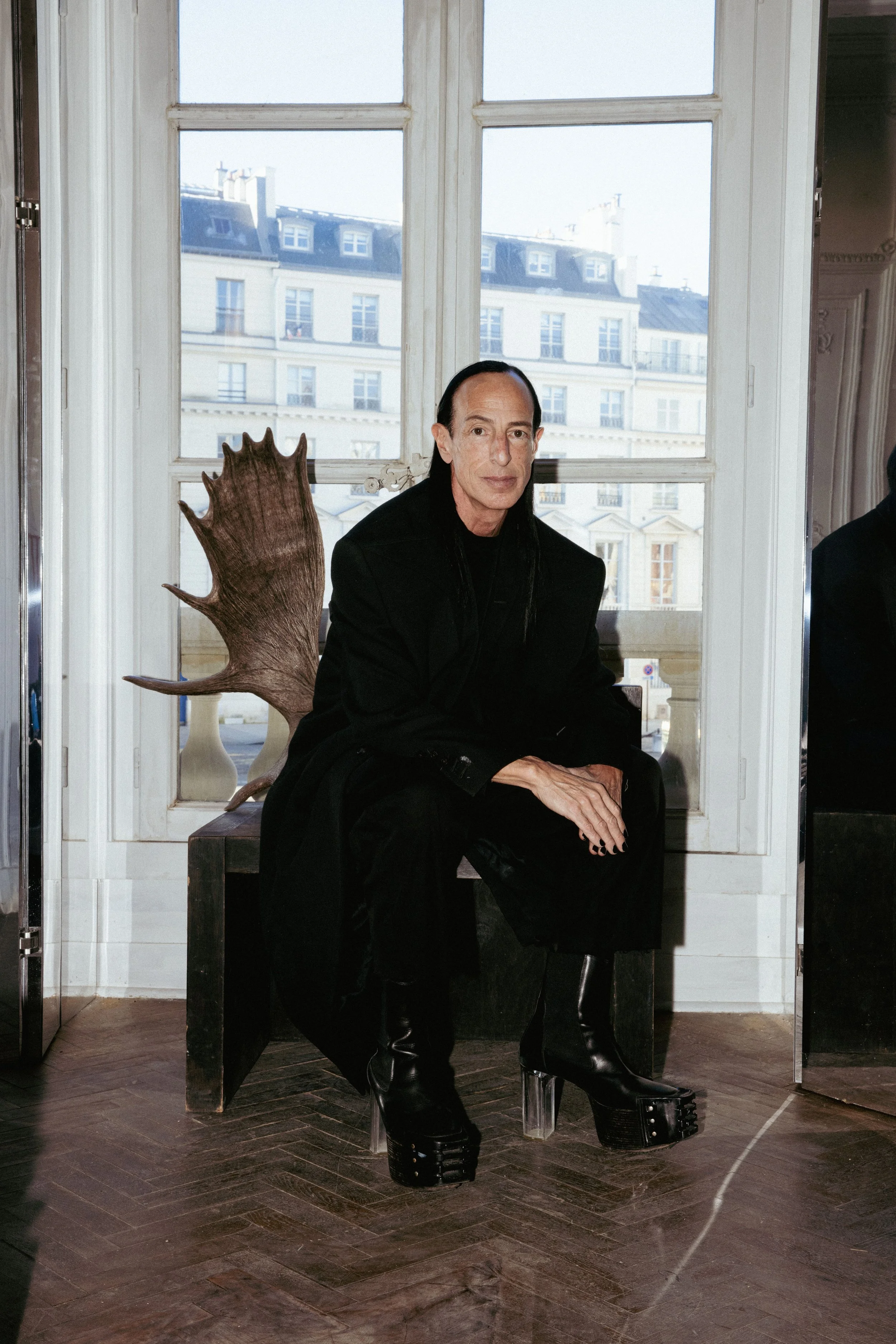 A person with long dark hair and pale skin, dressed in black, sitting on a wooden chair with a carved, spiked backrest. The person is wearing platform boots and black clothing. They are indoors near large windows showing Parisian buildings in the background.