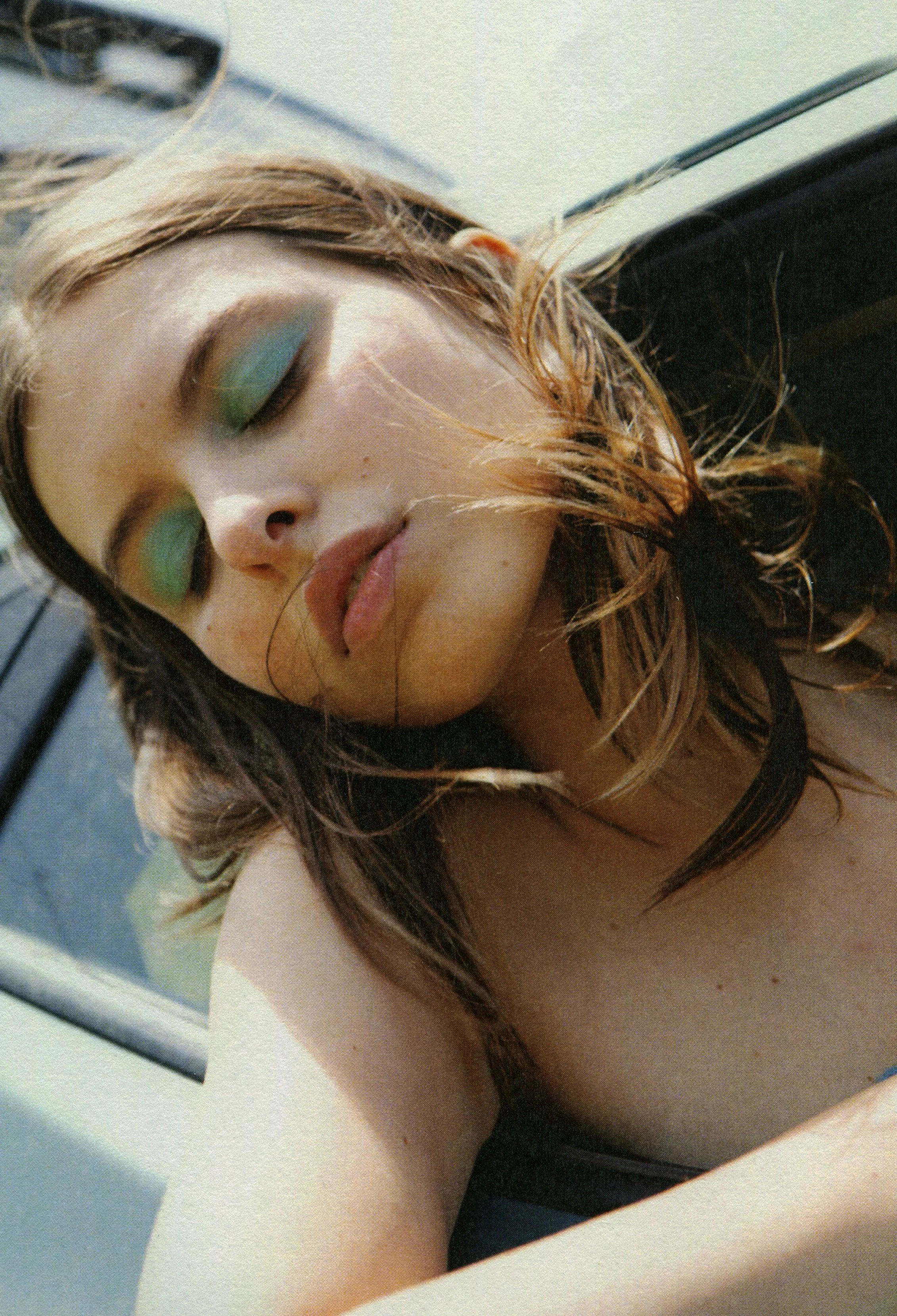 Close-up of a woman sleeping or resting her eyes, with makeup and wavy hair, on a white surface near a black object.